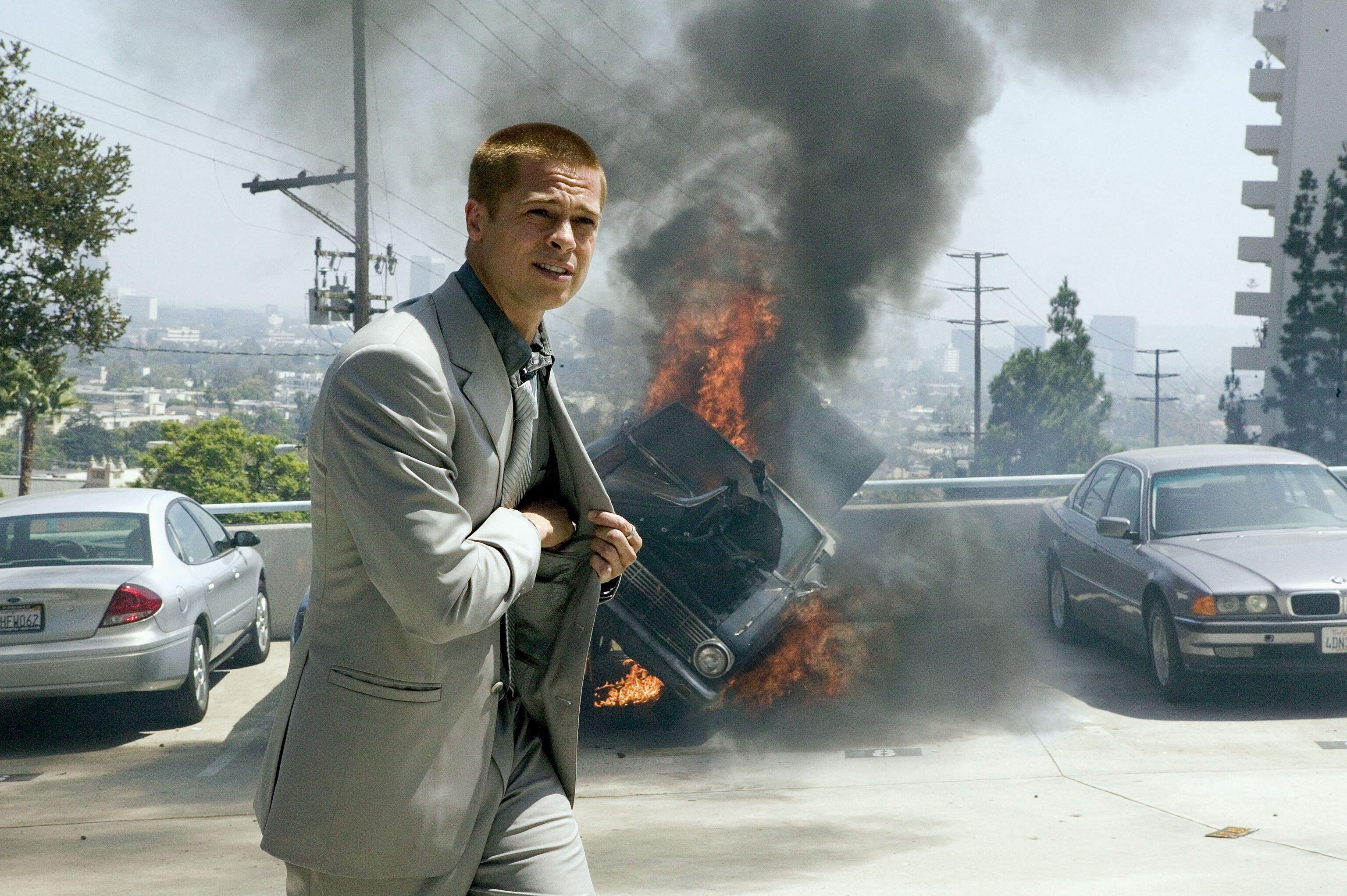 A man in a suit walks away from a burning car engulfed in flames and smoke in a parking lot.