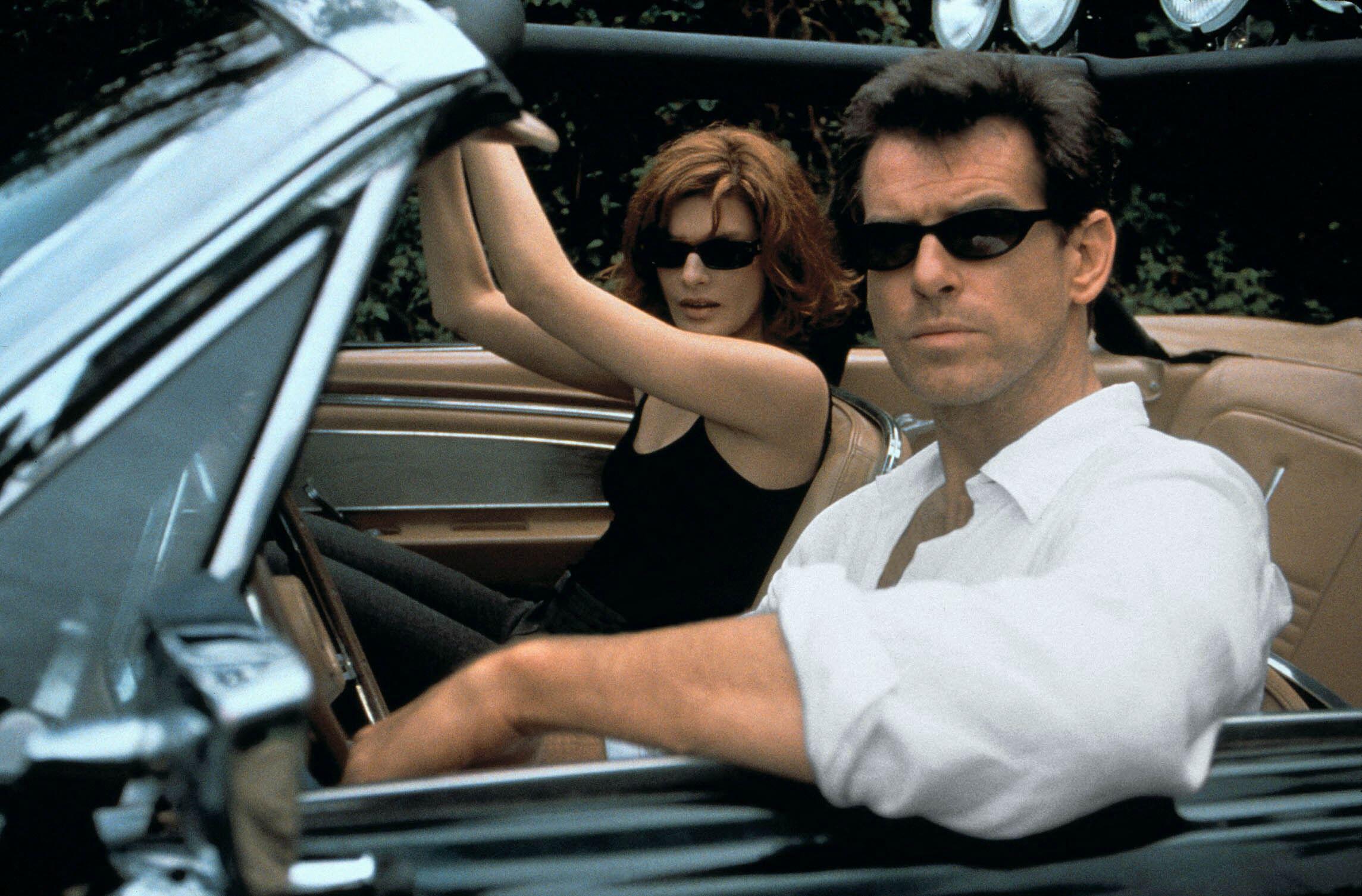 A man and a woman wearing sunglasses sit in a convertible car, looking serious.