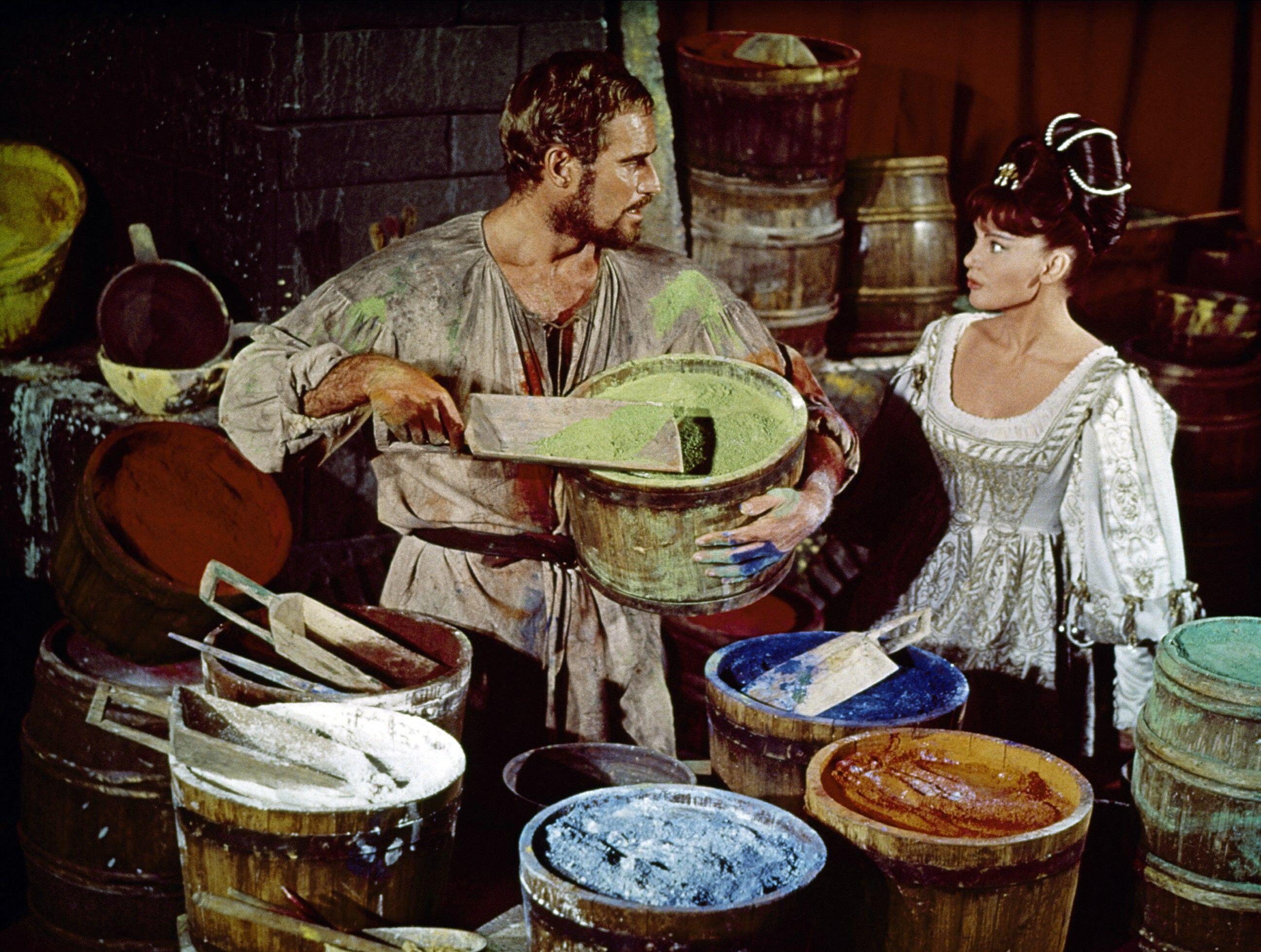 A man in rustic clothes mixes paint while a woman in a white dress stands beside him, surrounded by colorful buckets.