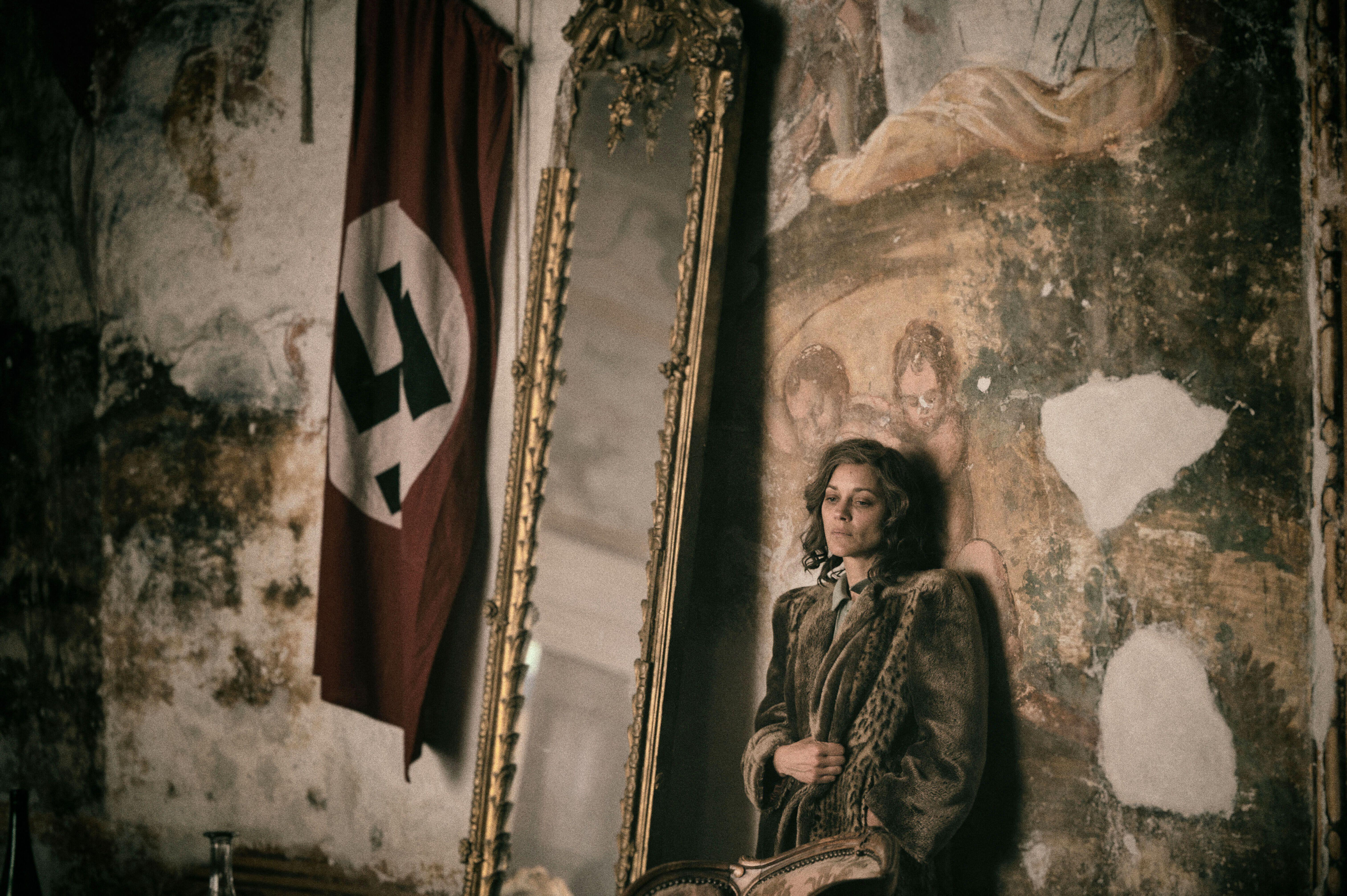 A woman in vintage clothing stands by a mirror and a Nazi flag in a worn, decorated room.