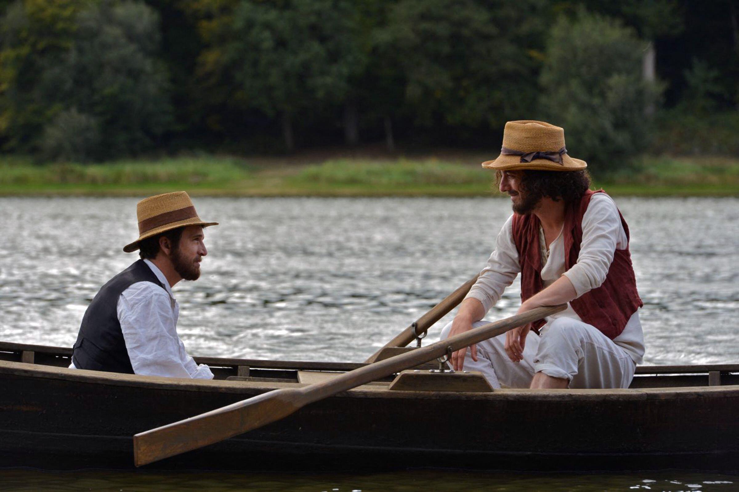 Two men in vintage clothing and straw hats sit in a wooden rowboat on a river.