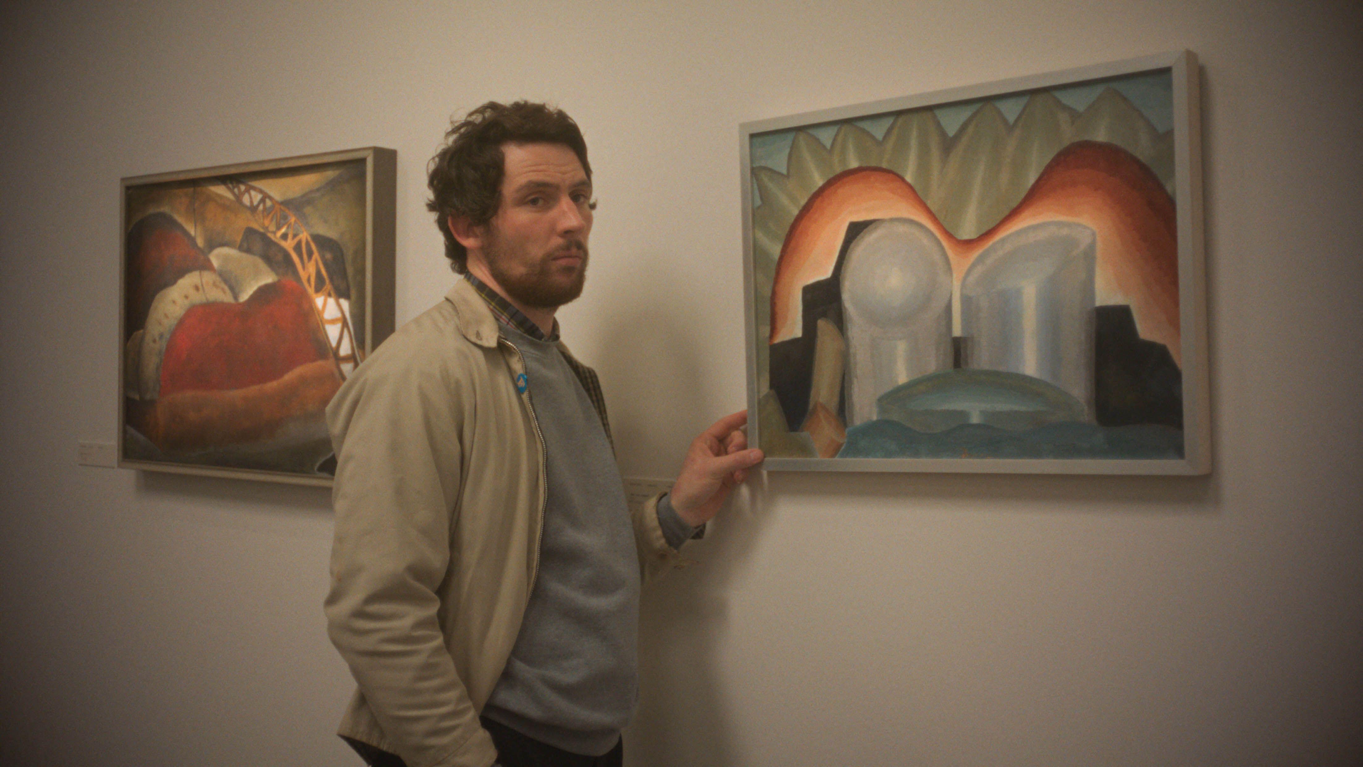 A man stands in an art gallery, pointing at a colorful abstract painting on the wall.