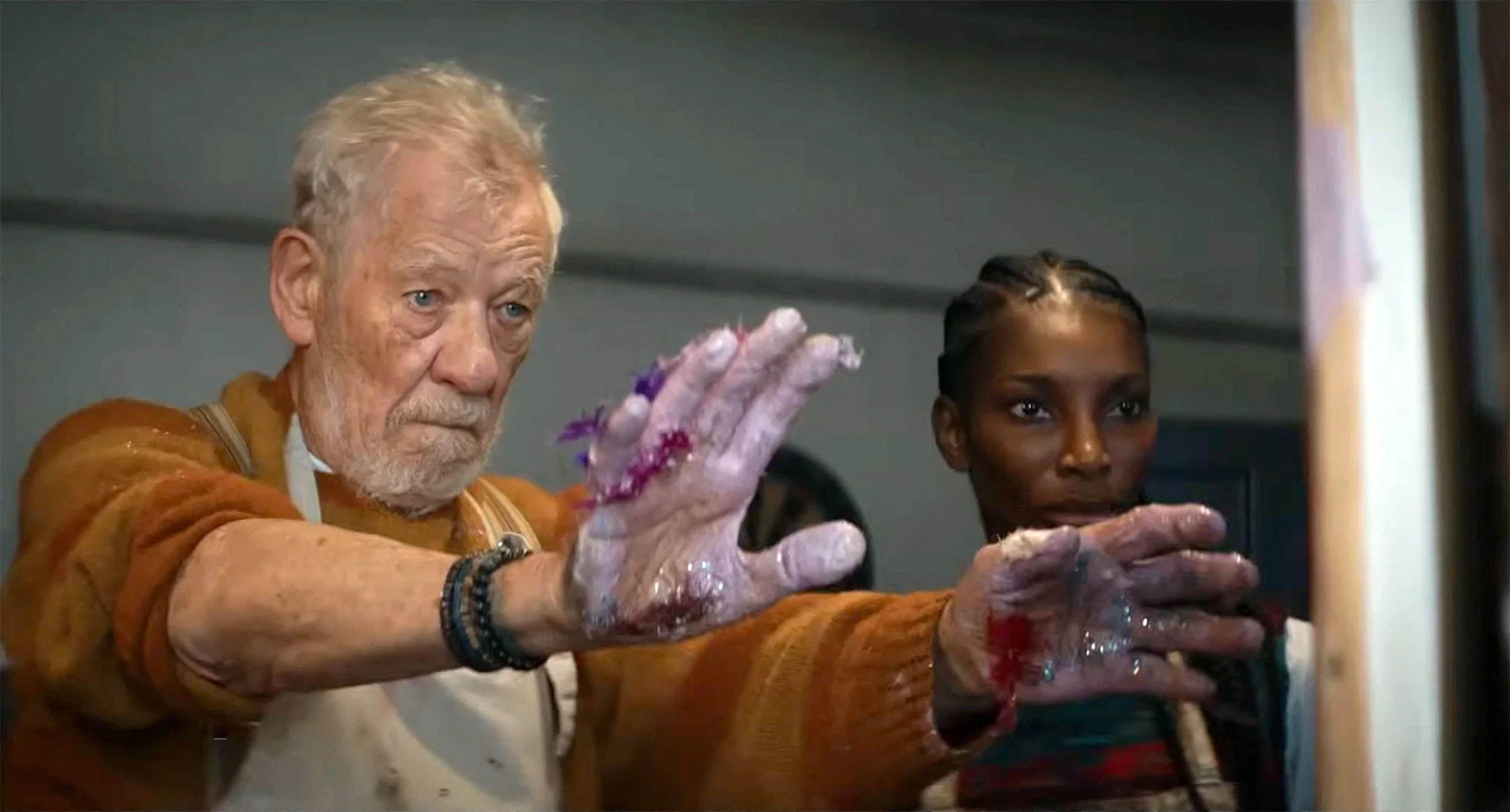 An elderly man with paint-covered hands stands beside a woman, both looking intently ahead.