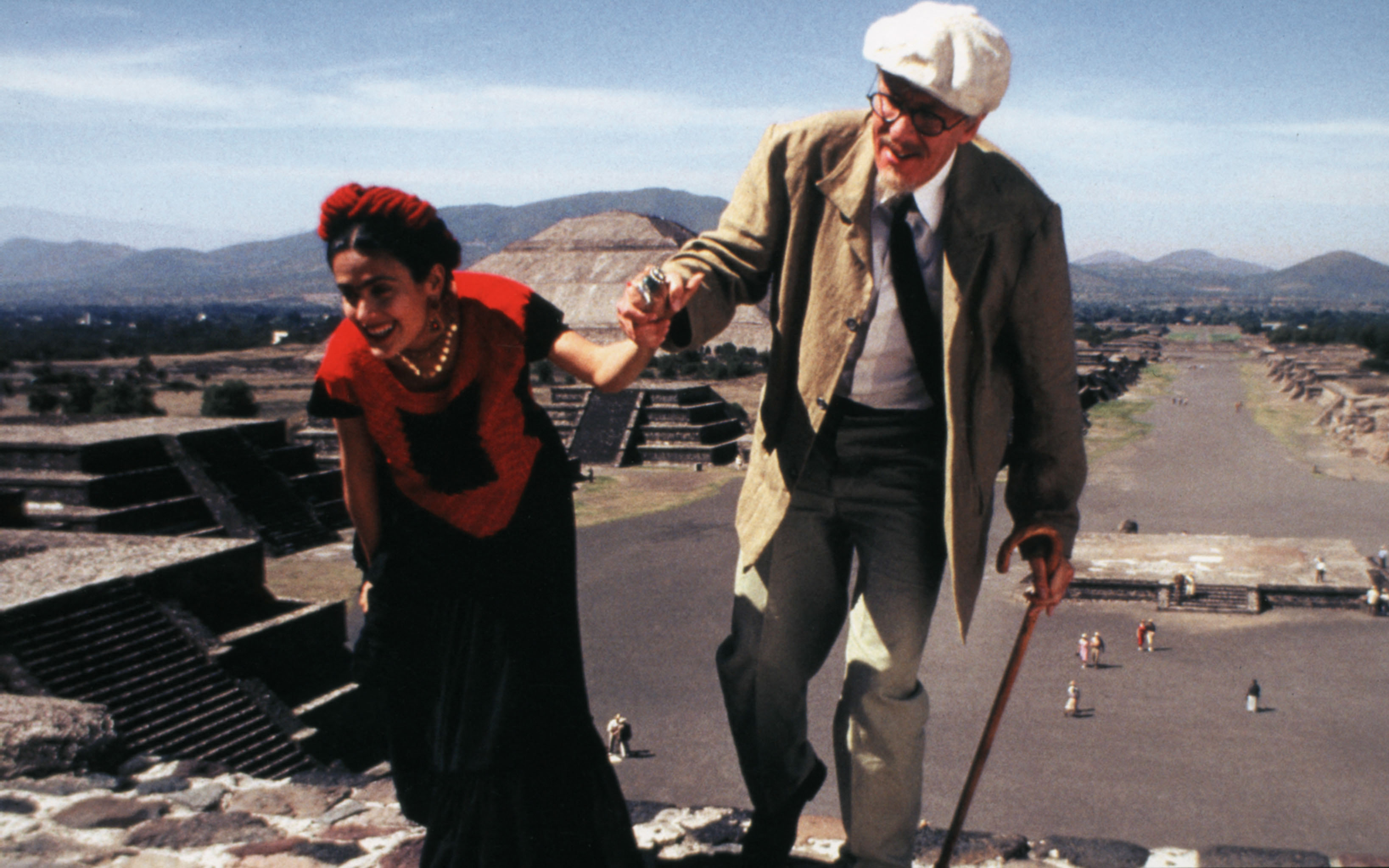 A woman in traditional dress helps an elderly man walk up ancient stone steps outdoors.