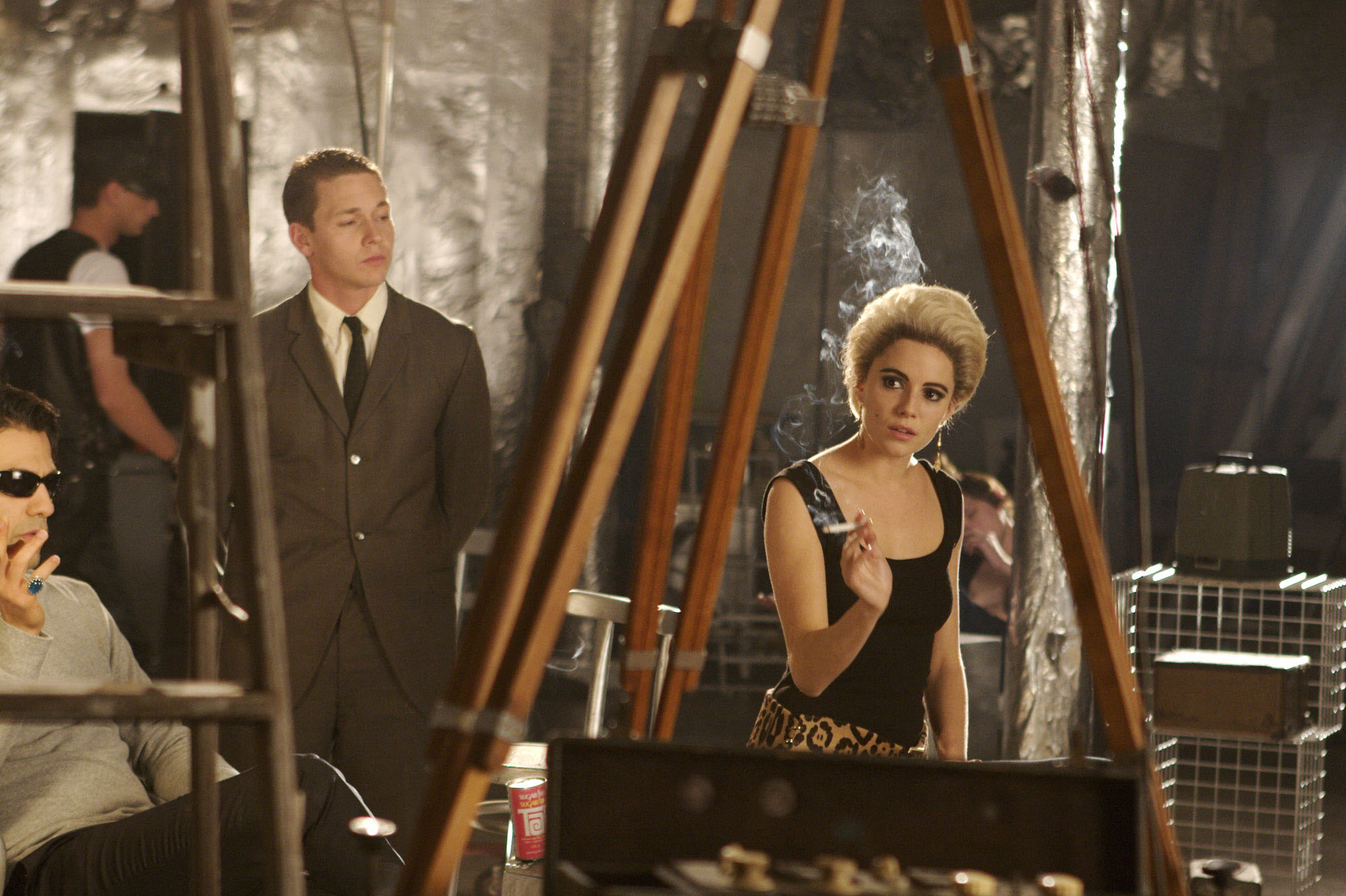 A woman with blonde hair holds a cigarette indoors, with two men and studio equipment around her.