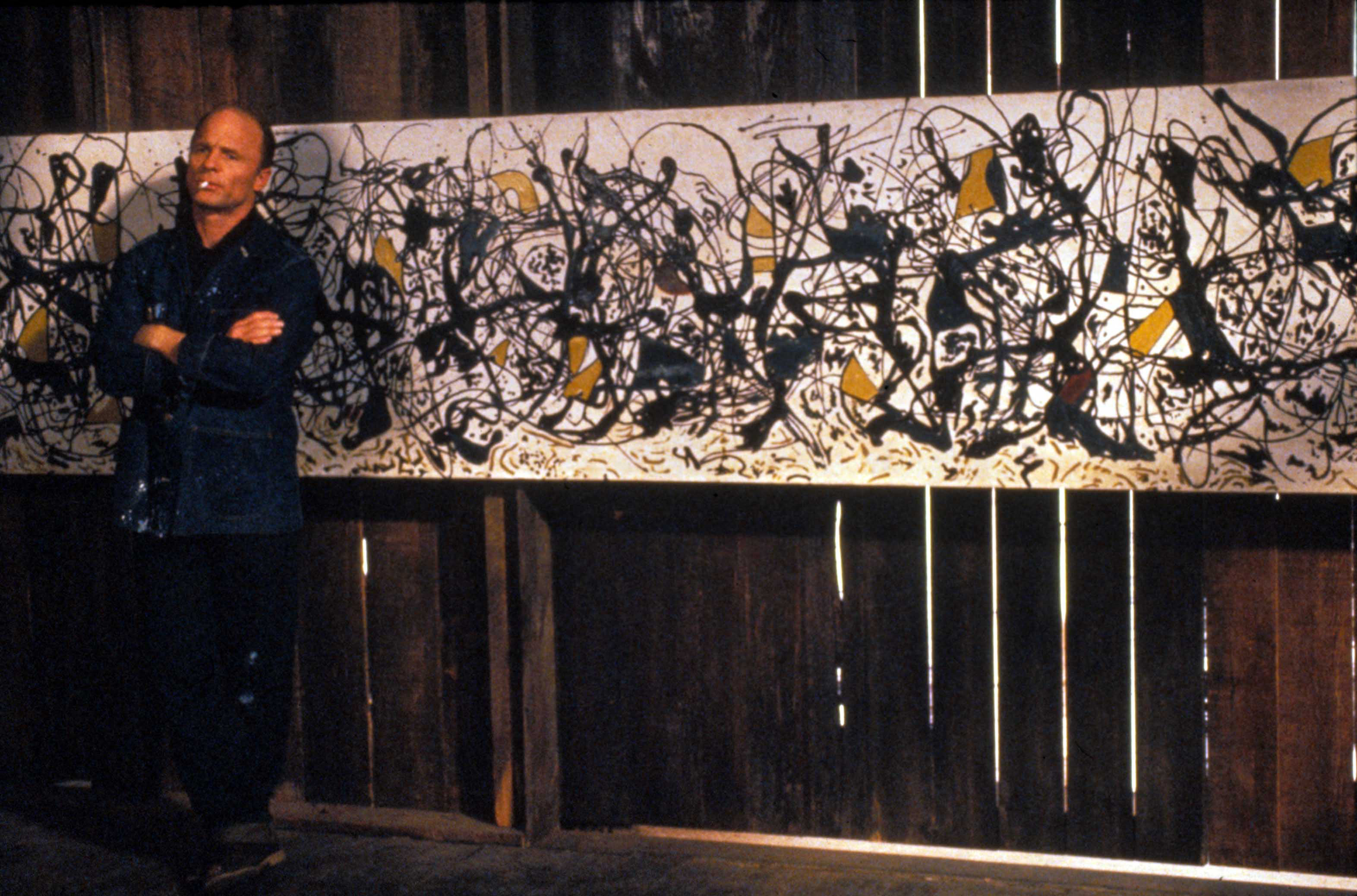 A man stands with arms crossed in front of a large abstract expressionist painting.