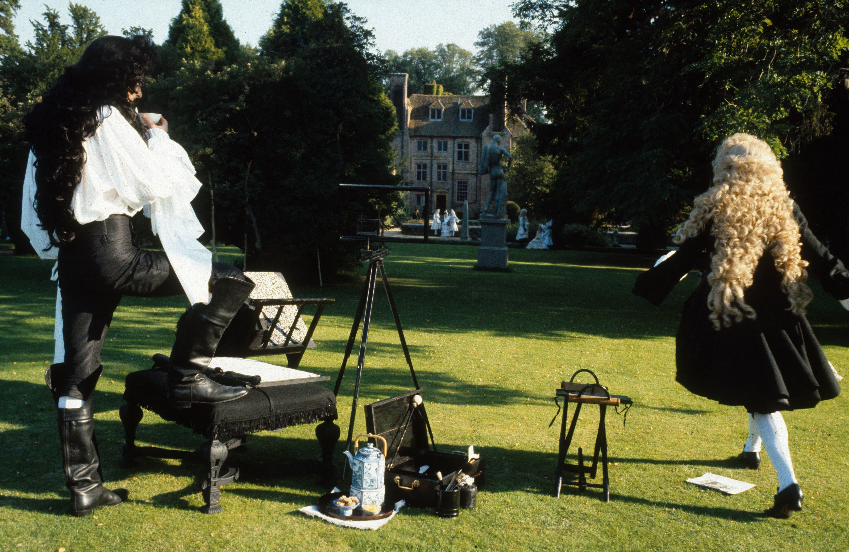 Two people in historical costumes stand on a lawn near art supplies, with a mansion in the background.