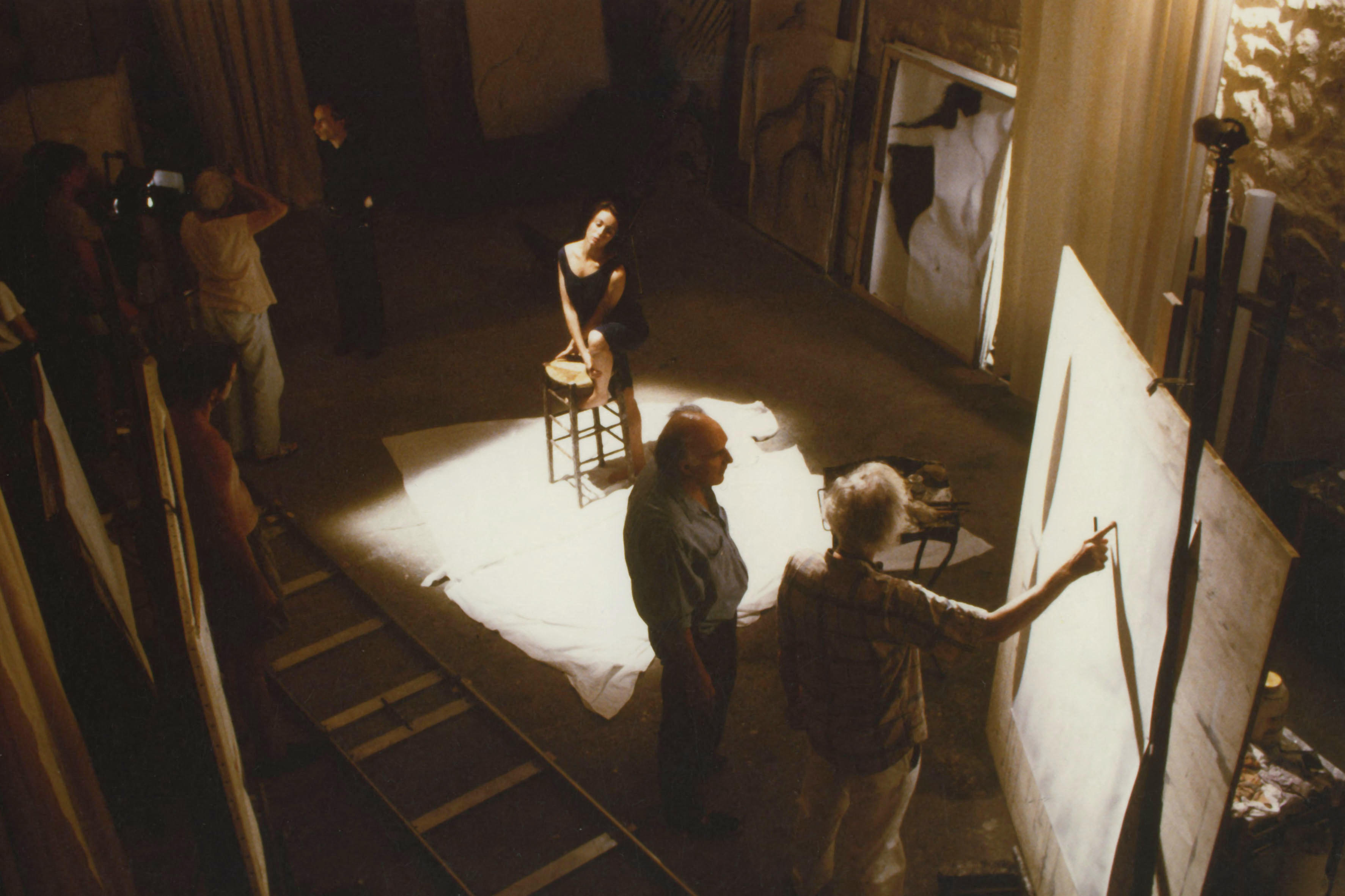 A group of people observe as an artist sketches a woman posing on a stool under a spotlight.