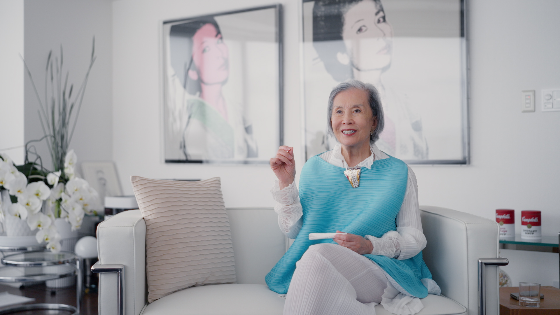 An elderly woman sits on a modern white chair in a bright room with art and flowers behind her.