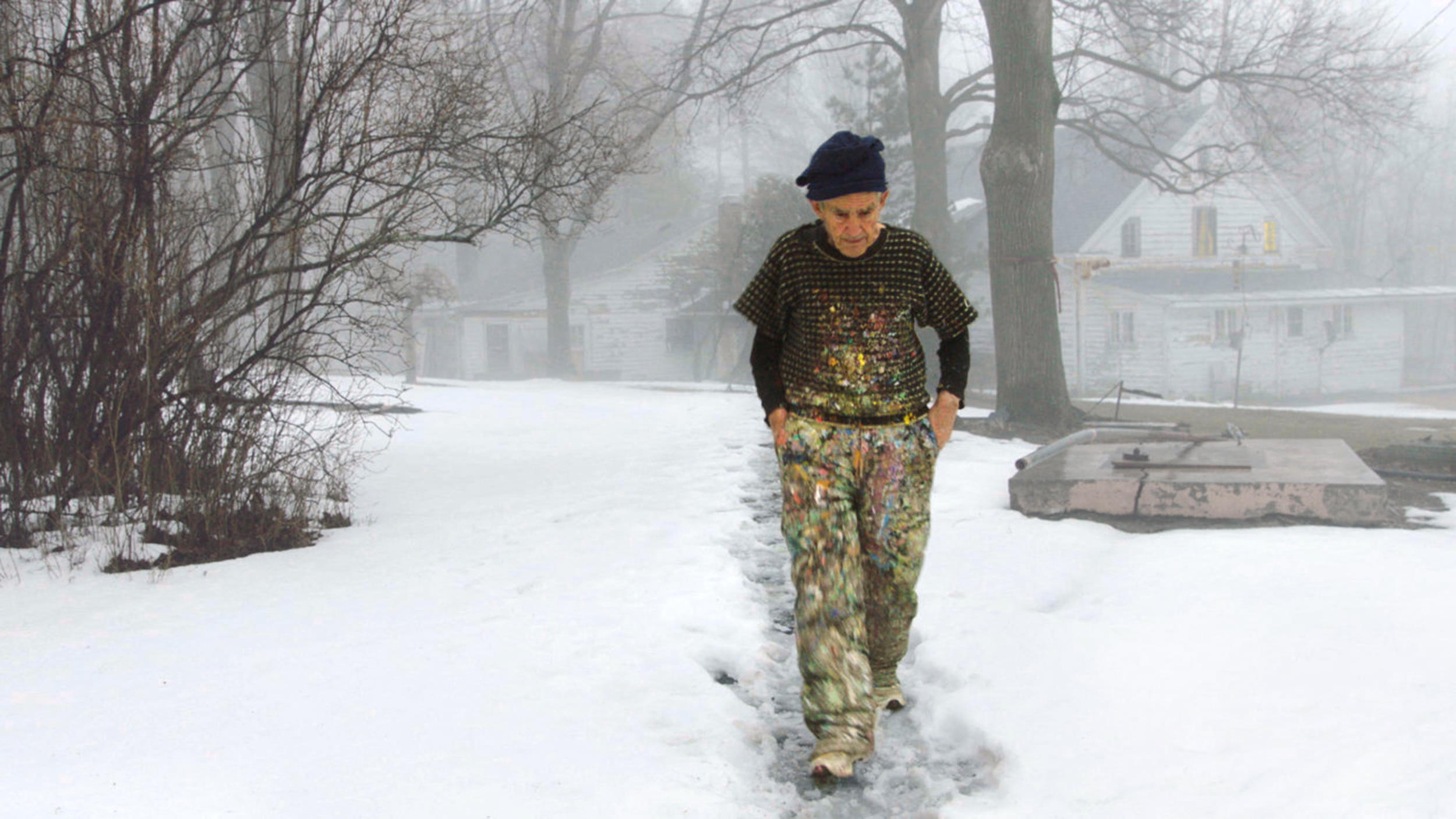 An elderly person in paint-splattered clothes walks on a snowy path in a foggy outdoor setting.