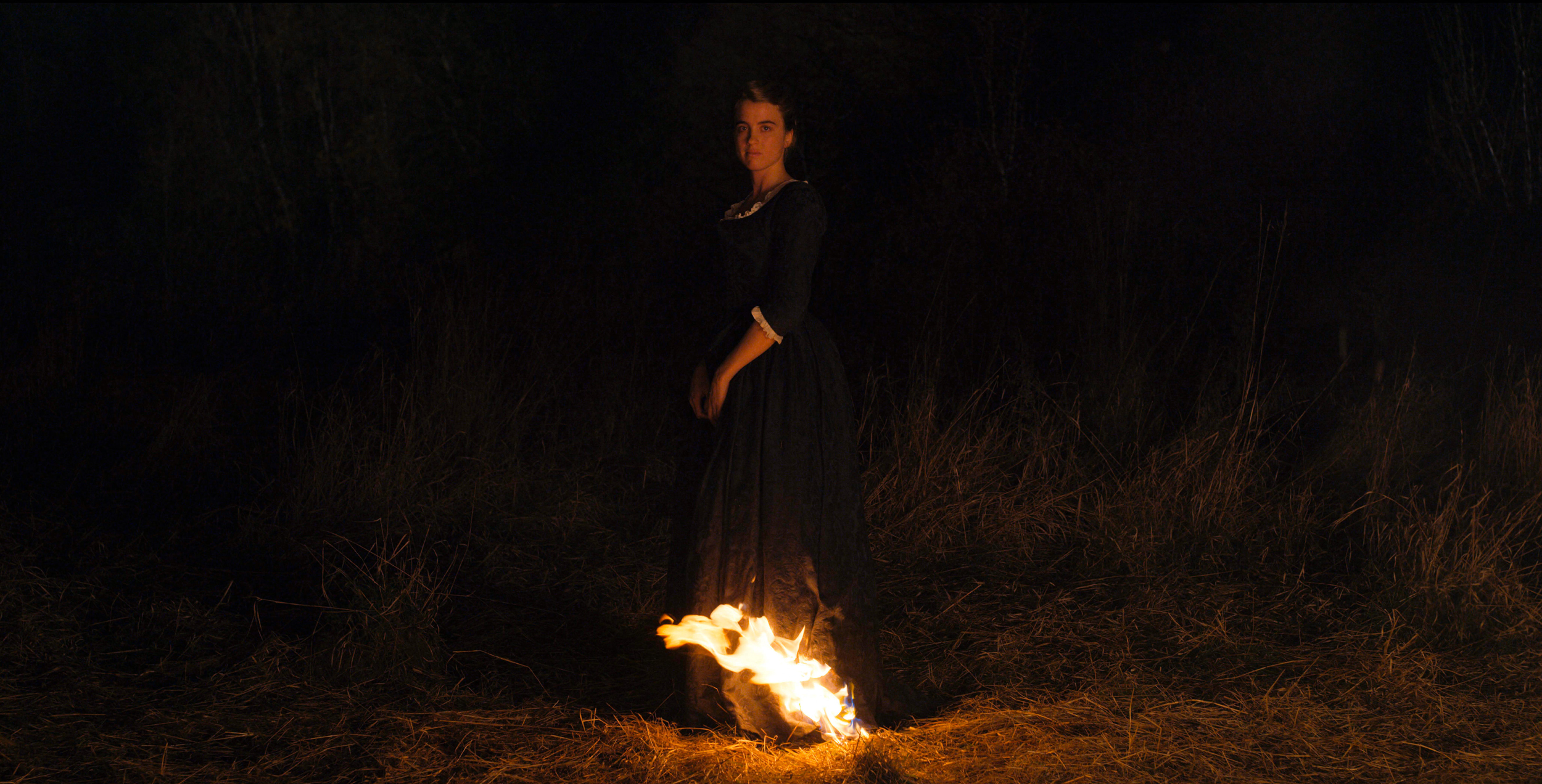 A woman in a dark dress stands in a field at night with fire at the hem of her dress.