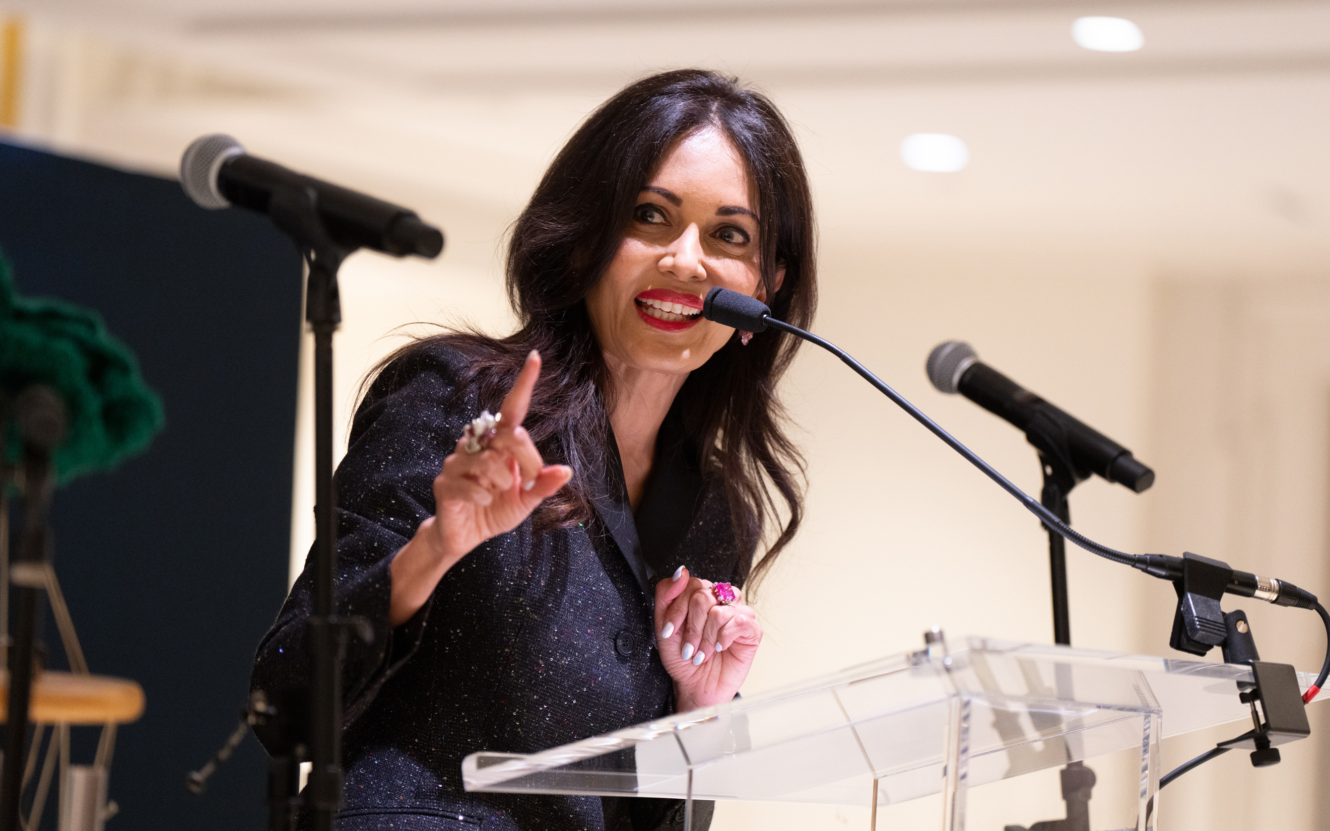 A woman is speaking and gesturing at a podium with microphones in a bright indoor setting.
