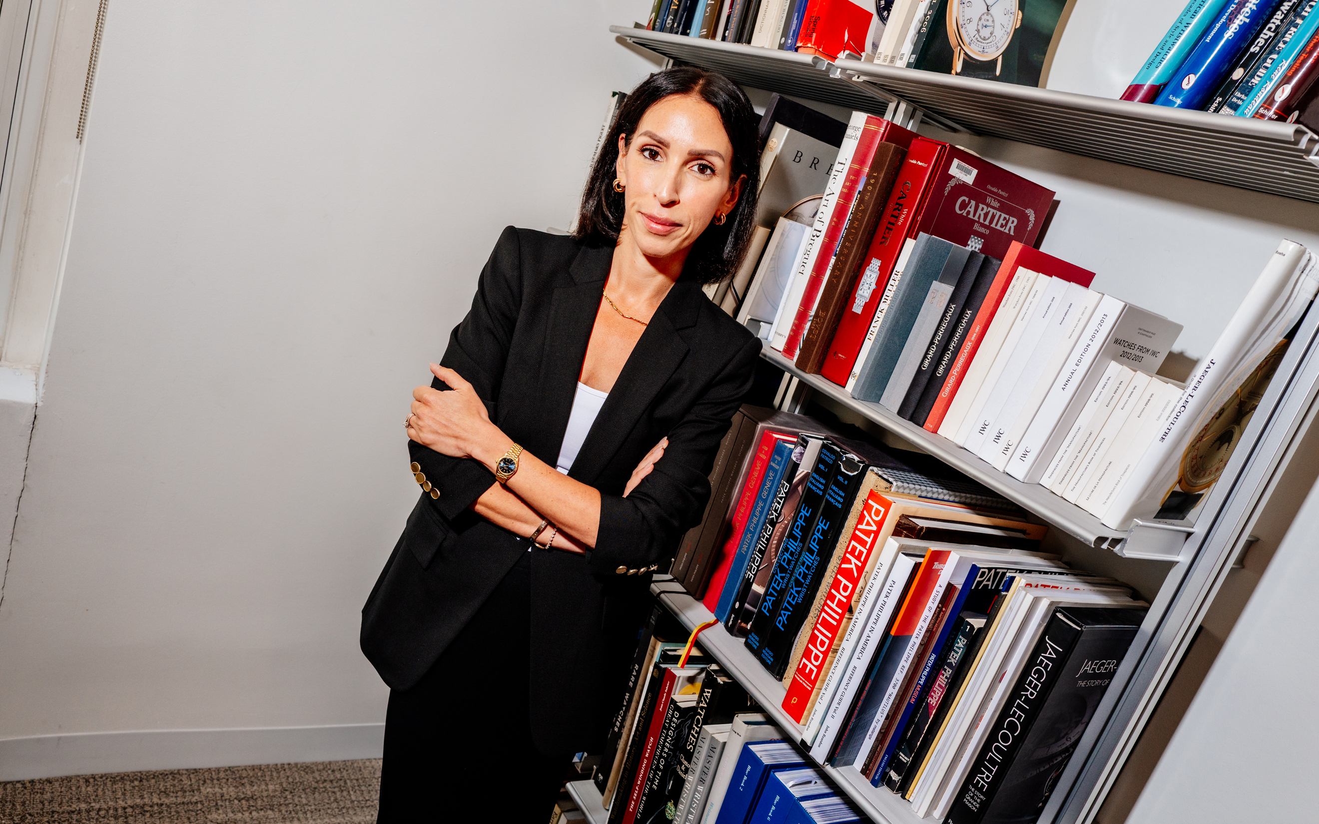 A woman in a black blazer stands beside a bookshelf filled with books and magazines.