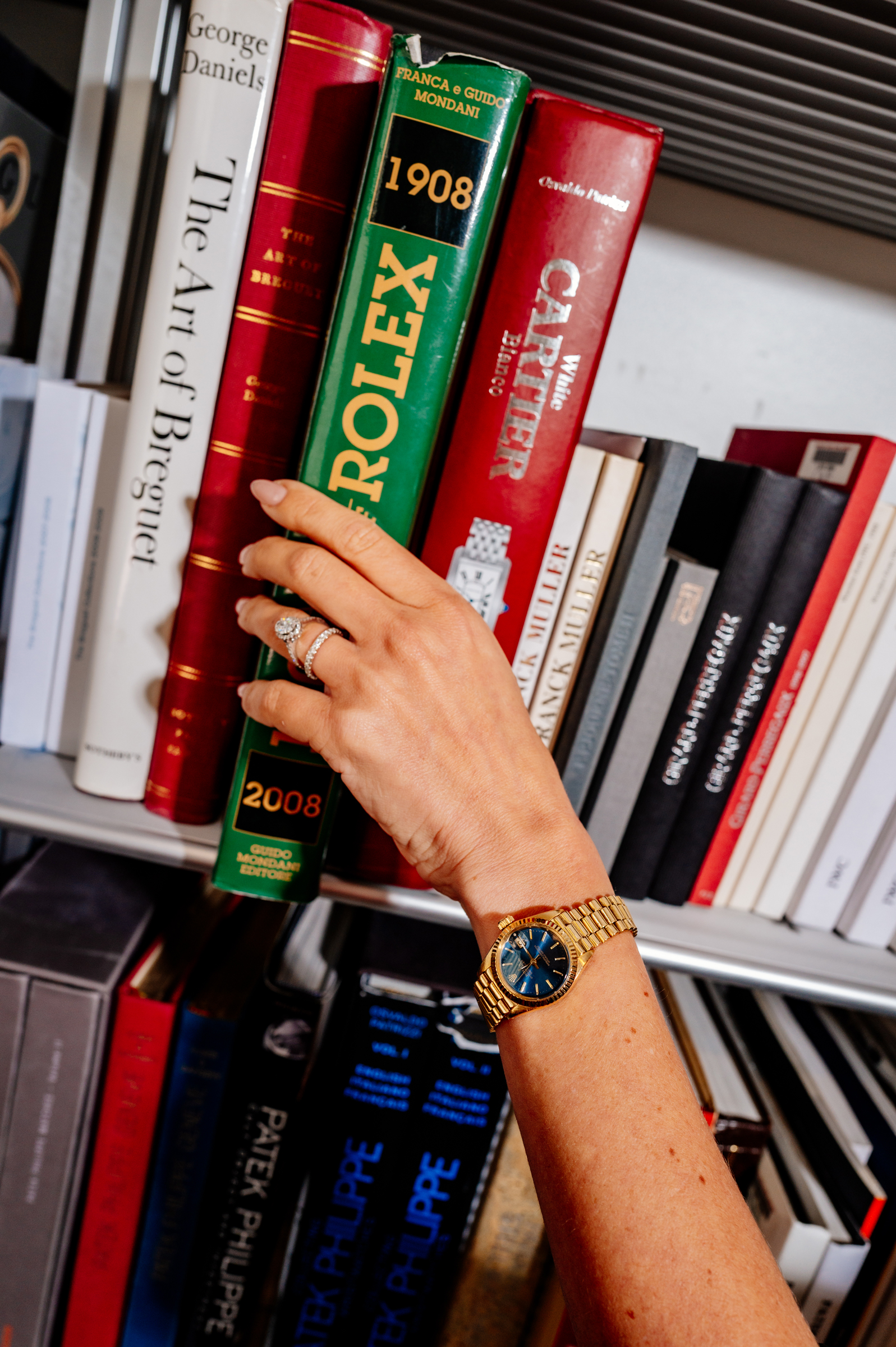 A hand wearing a gold watch is reaching for a green Rolex book on a bookshelf.