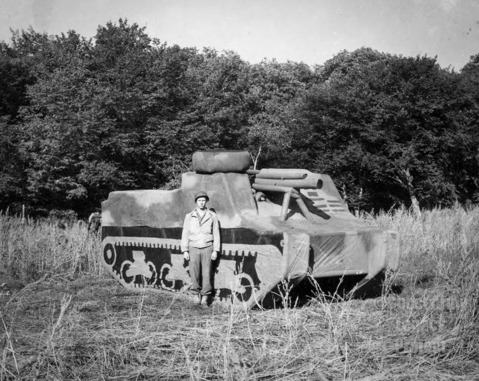 A man stands next to a fake tank made of canvas in a grassy field with trees in the background.