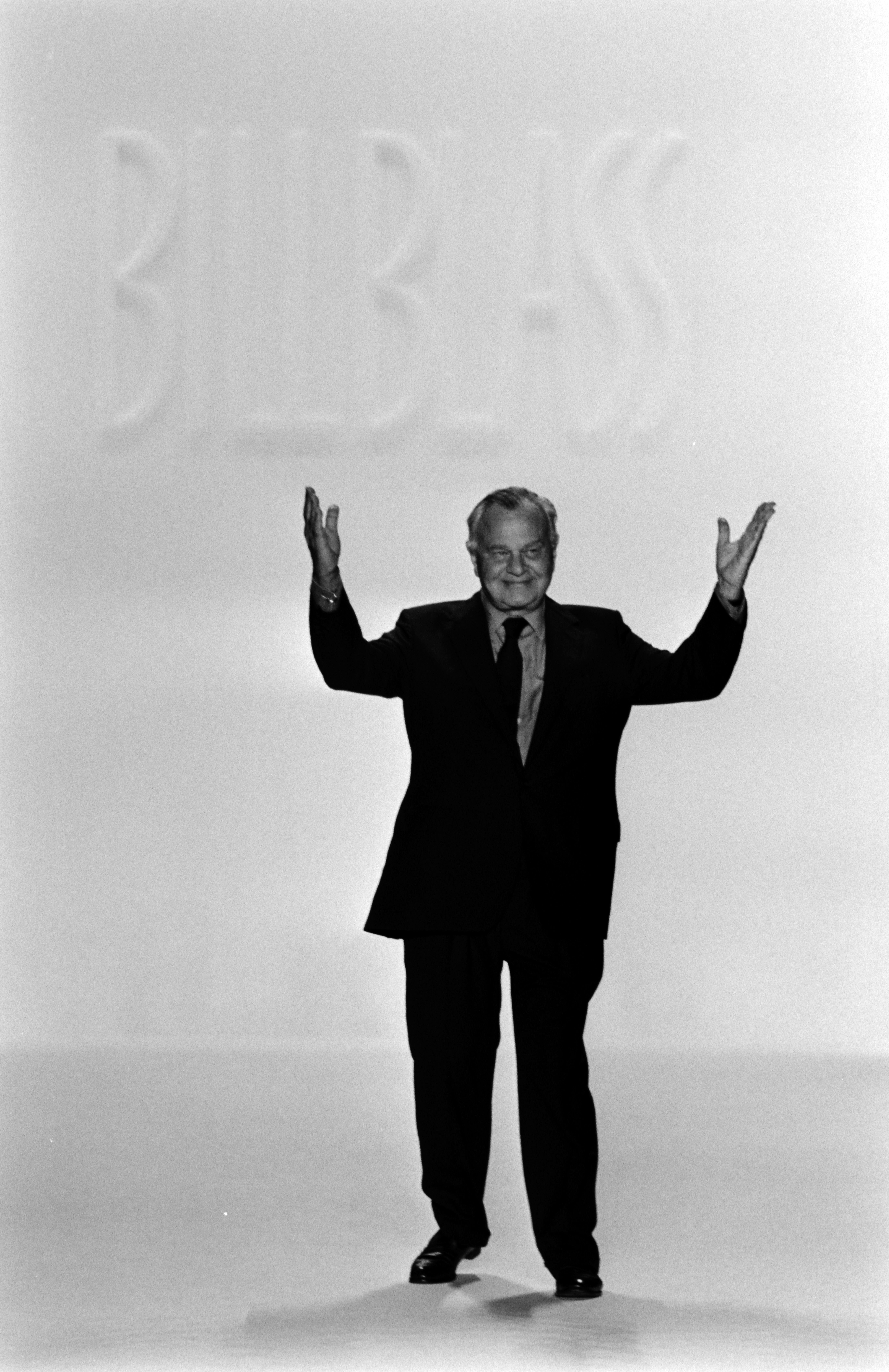 A man in a suit stands on a runway with his arms raised, with "BILL BLASS" in the background.