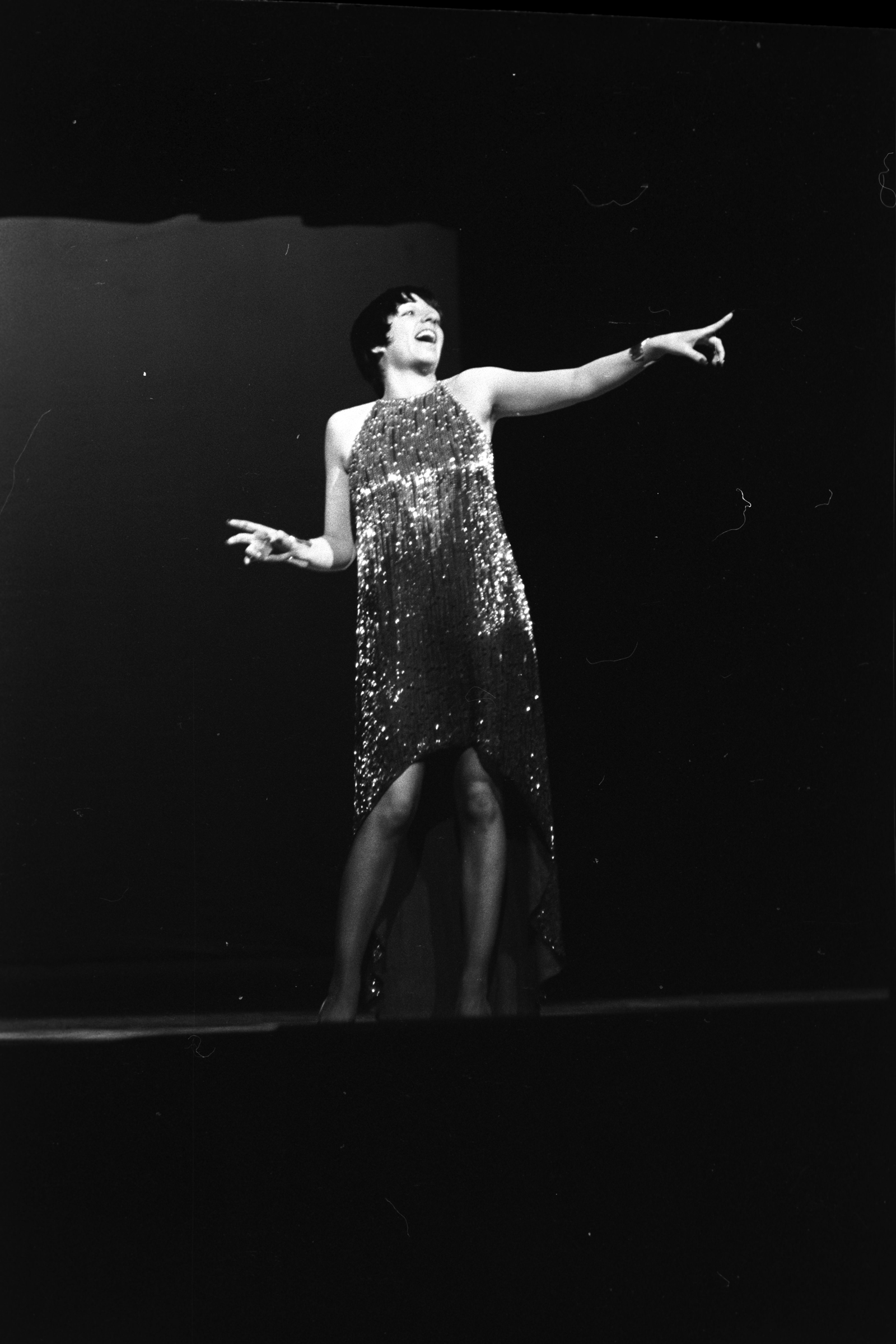 A performer in a sequined dress is singing and gesturing energetically on stage.