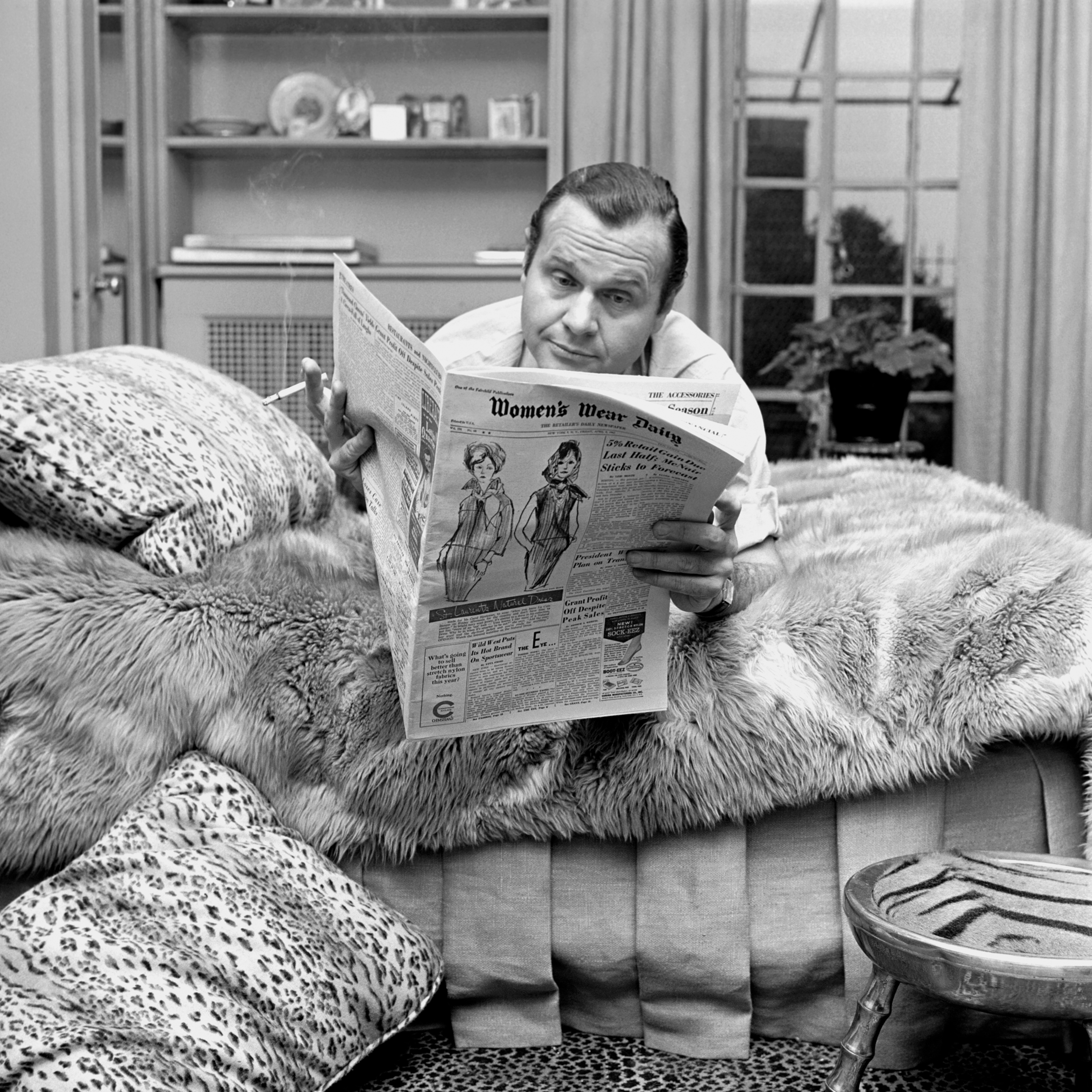 A man on a fur-covered bed reads a newspaper and holds a cigarette in a stylish room.