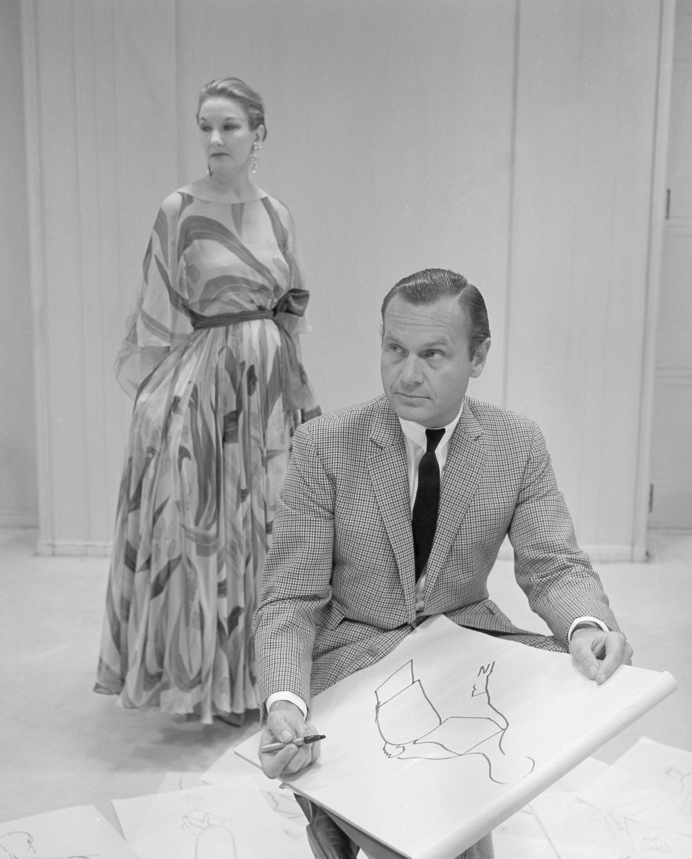 A man sketches on a large pad while a woman in a flowing dress stands behind him.