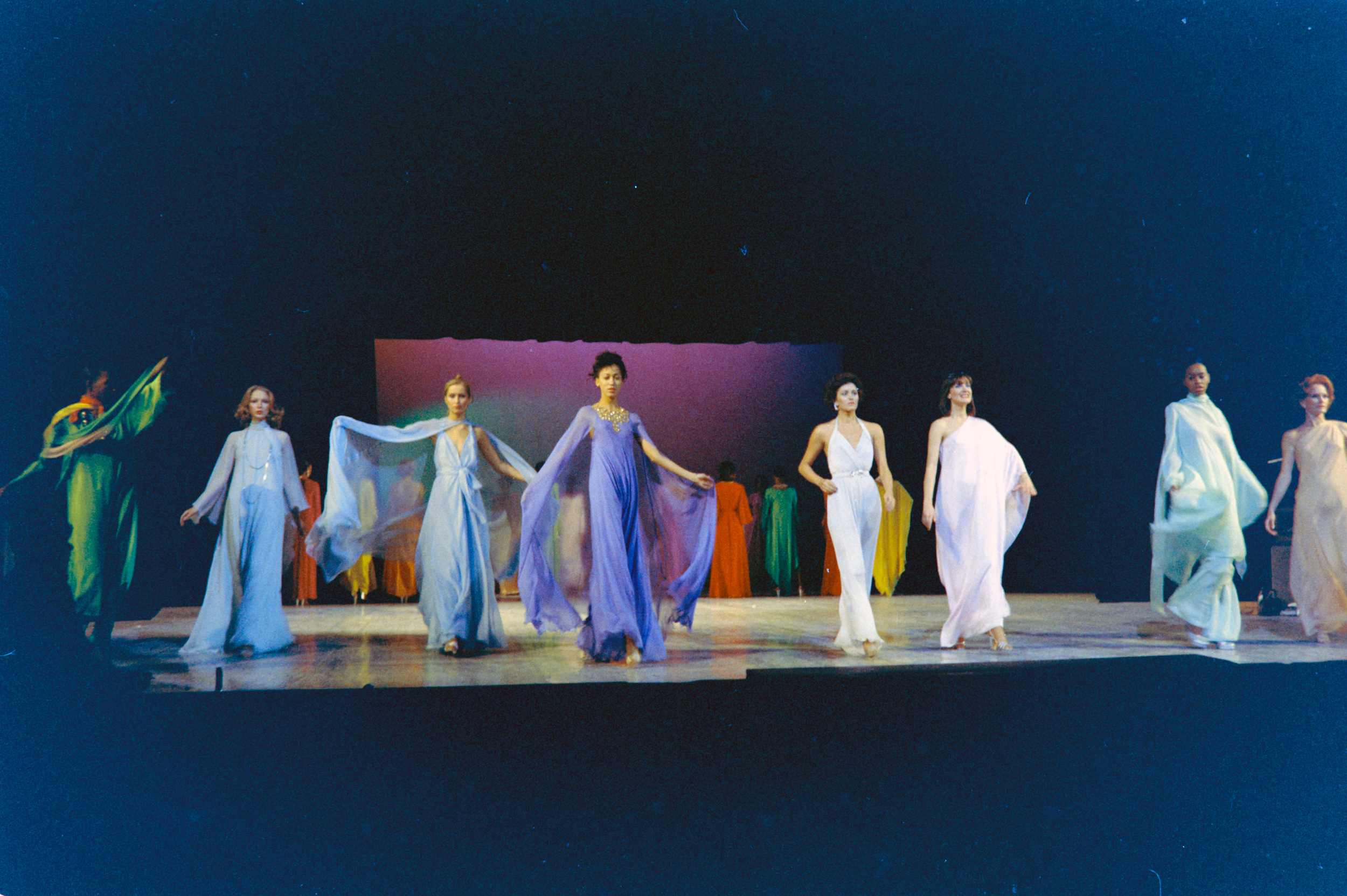 A group of models walk on a runway wearing flowing, colorful dresses under stage lights.