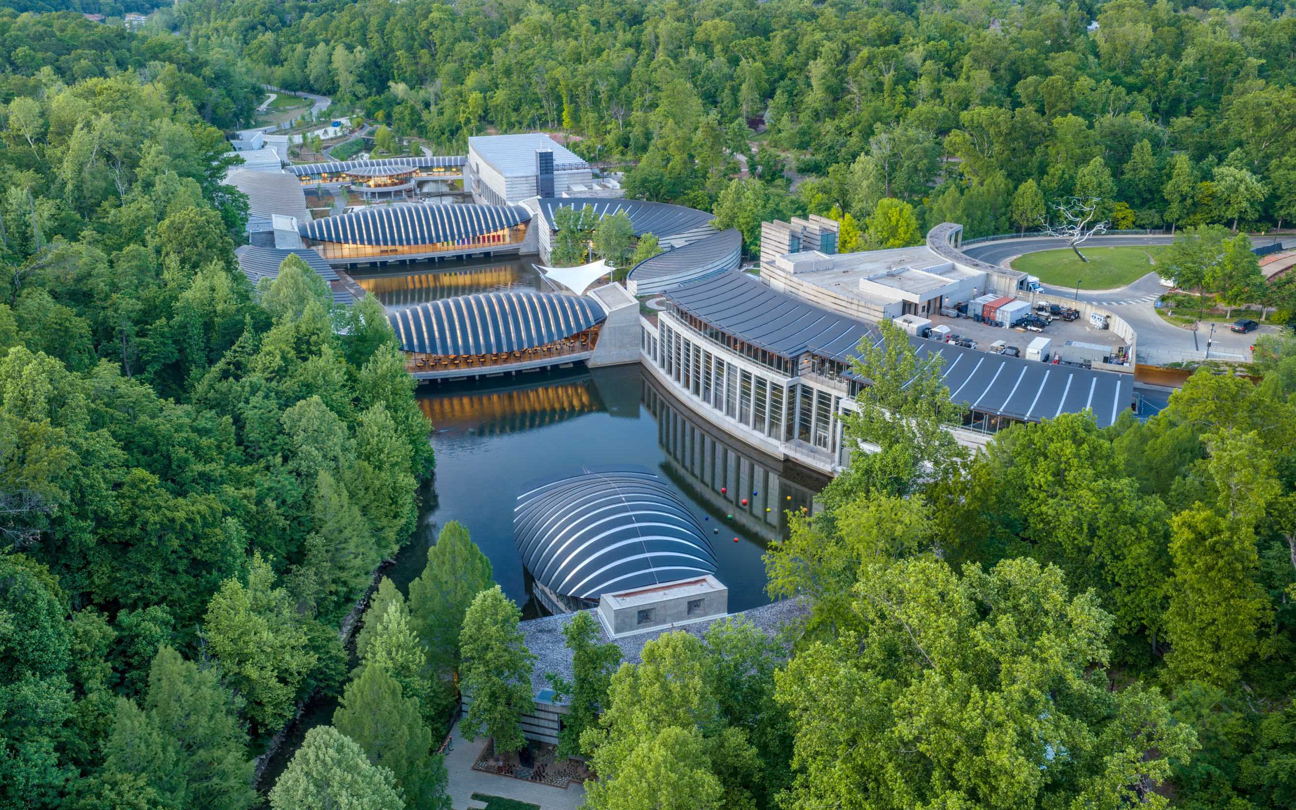 A modern, uniquely designed building complex surrounded by dense green forest and water features.