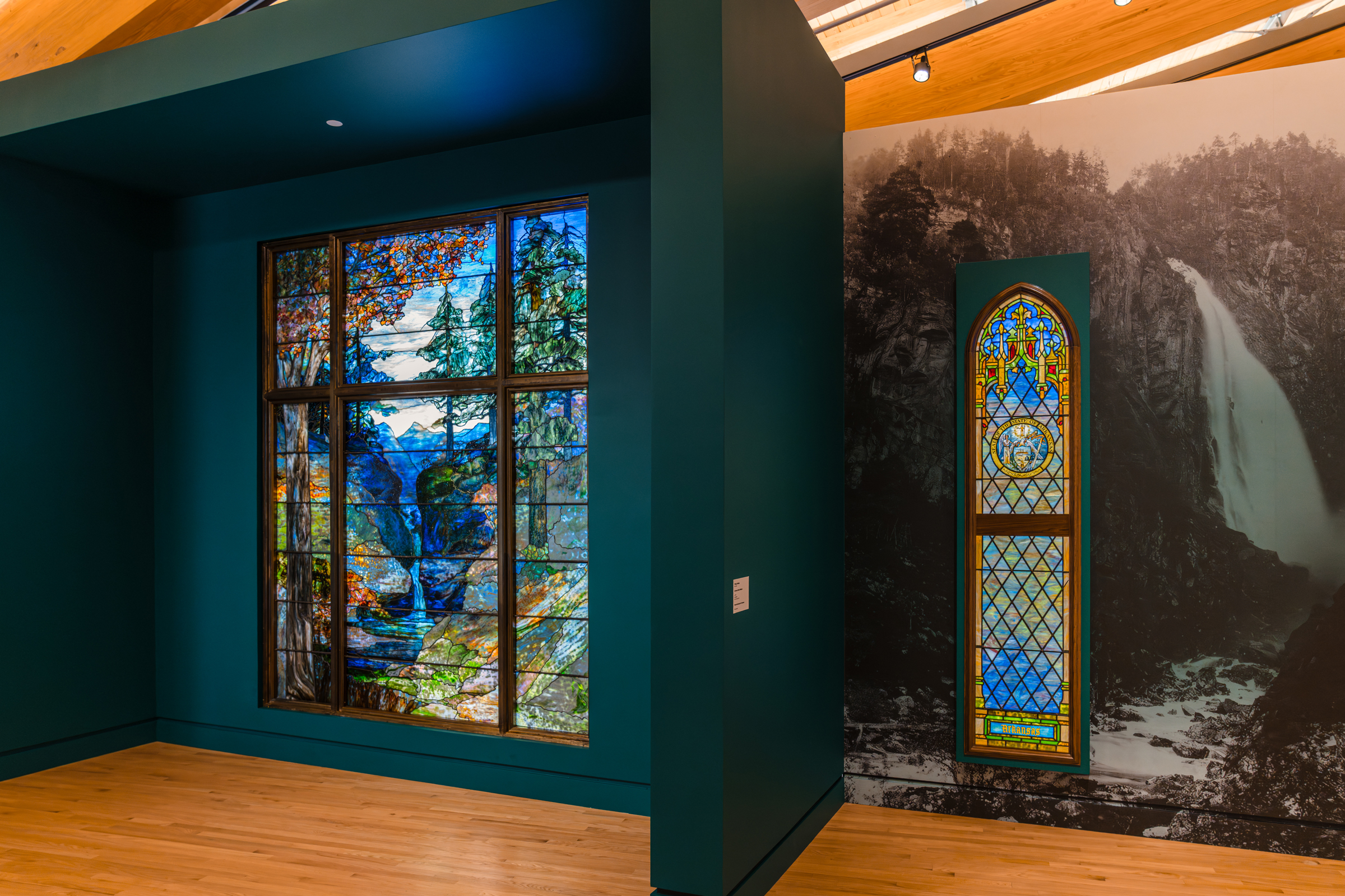 Two stained glass windows are displayed in a museum setting with one depicting a landscape scene.