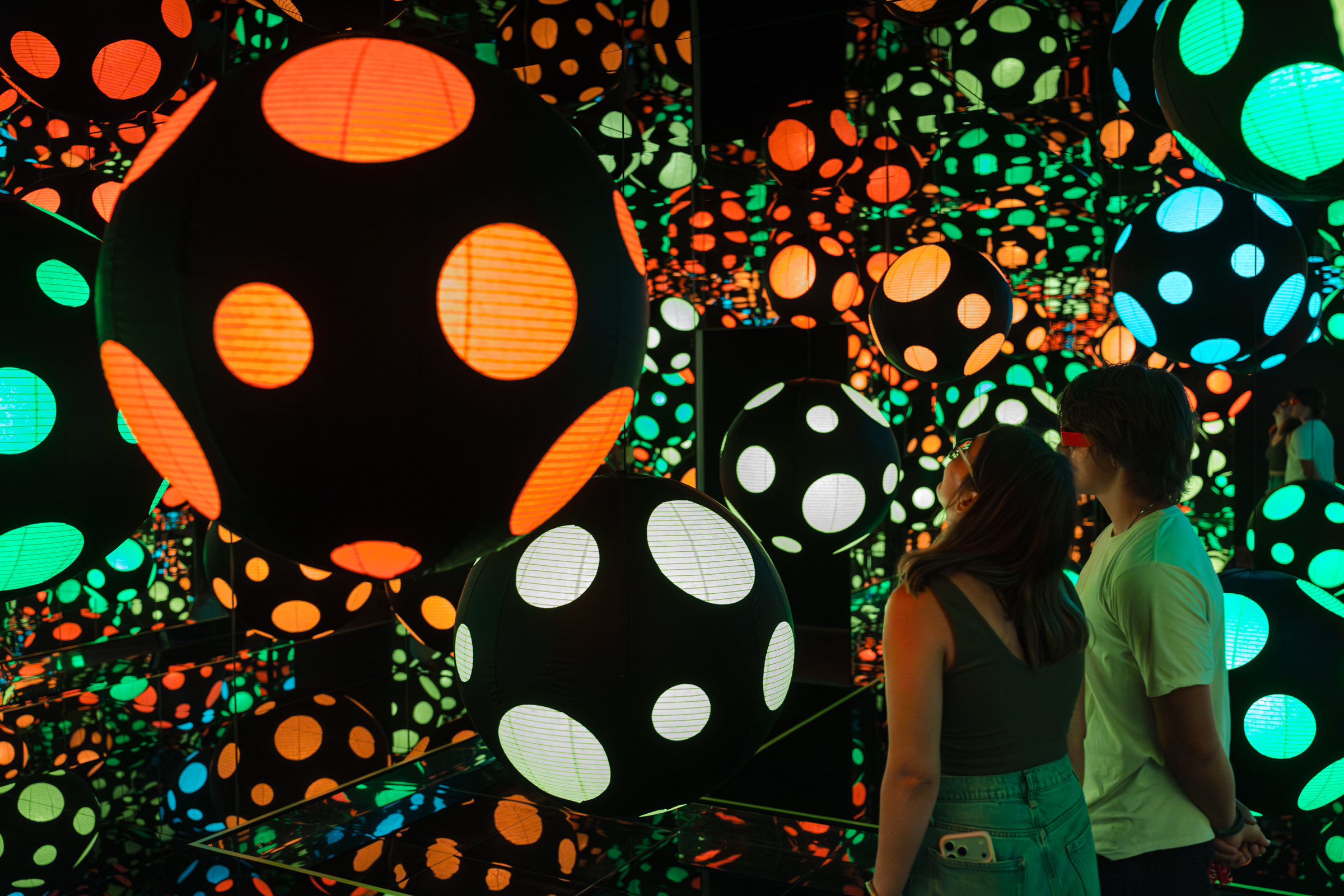 Two people observe a colorful, immersive art installation filled with glowing polka-dot spheres.