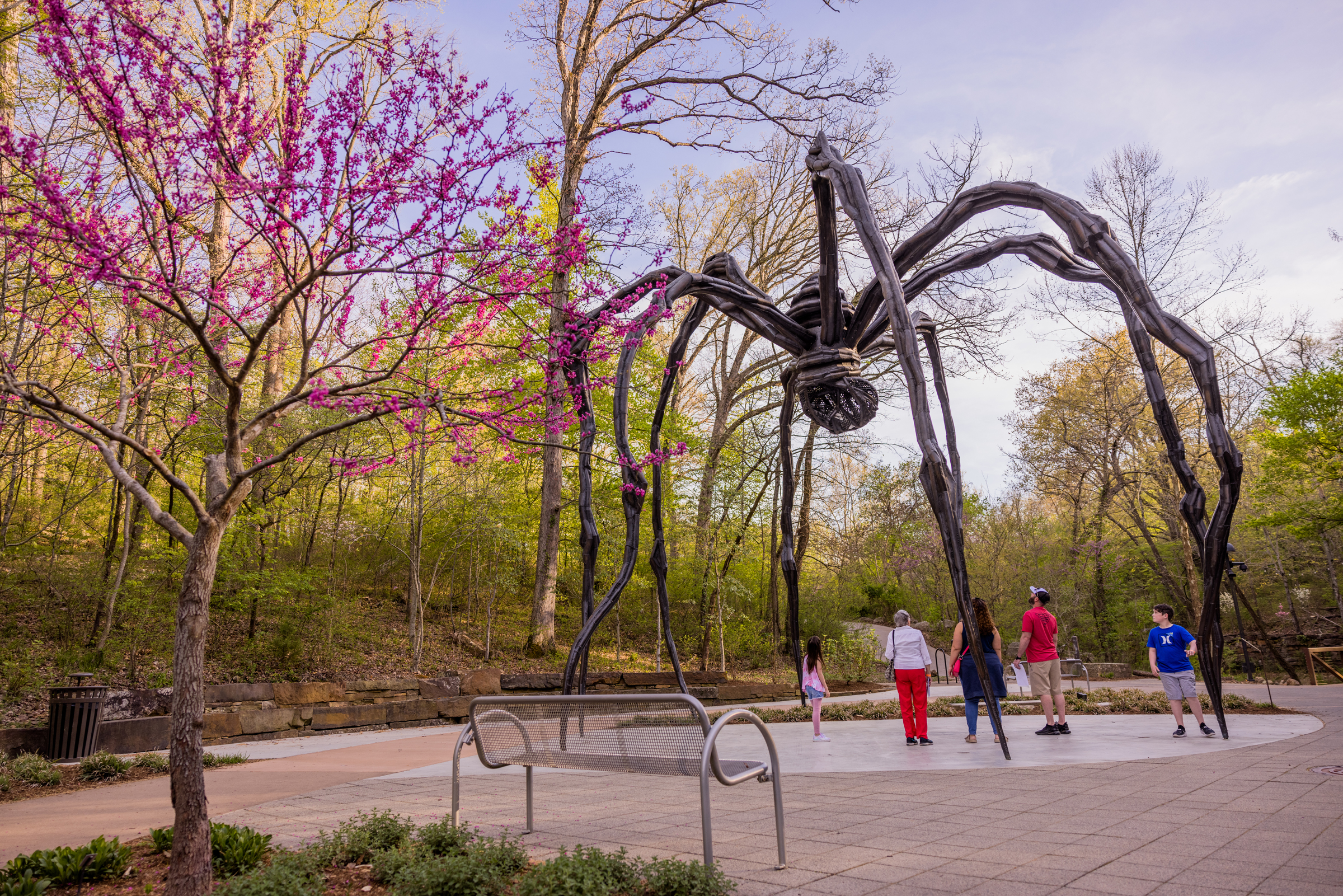 People look at a giant spider sculpture in a park surrounded by trees and blooming flowers.