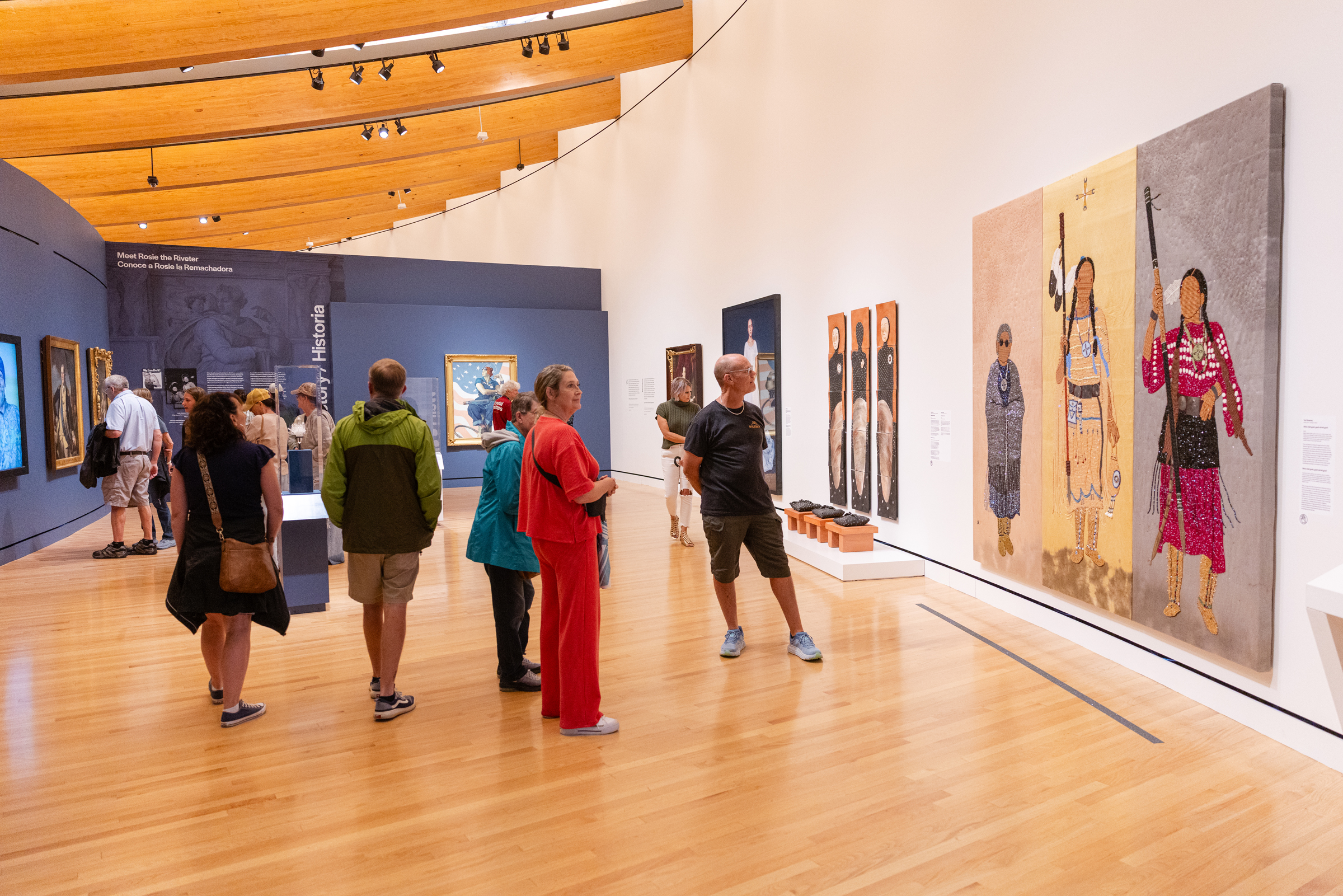 People are viewing various artworks in a spacious, modern art gallery with wooden floors.