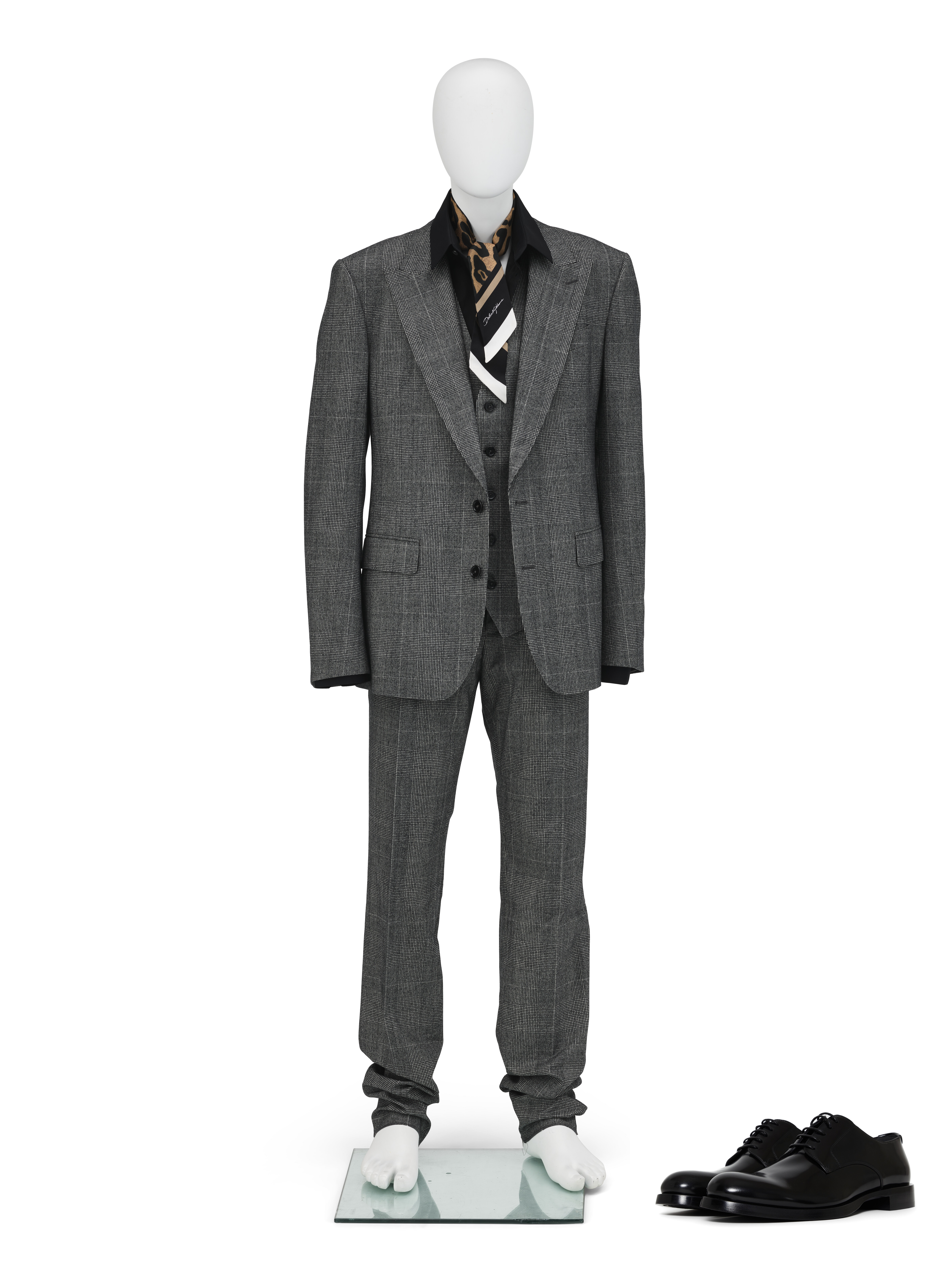 A mannequin is dressed in a gray suit with a black shirt and patterned scarf, next to black shoes.