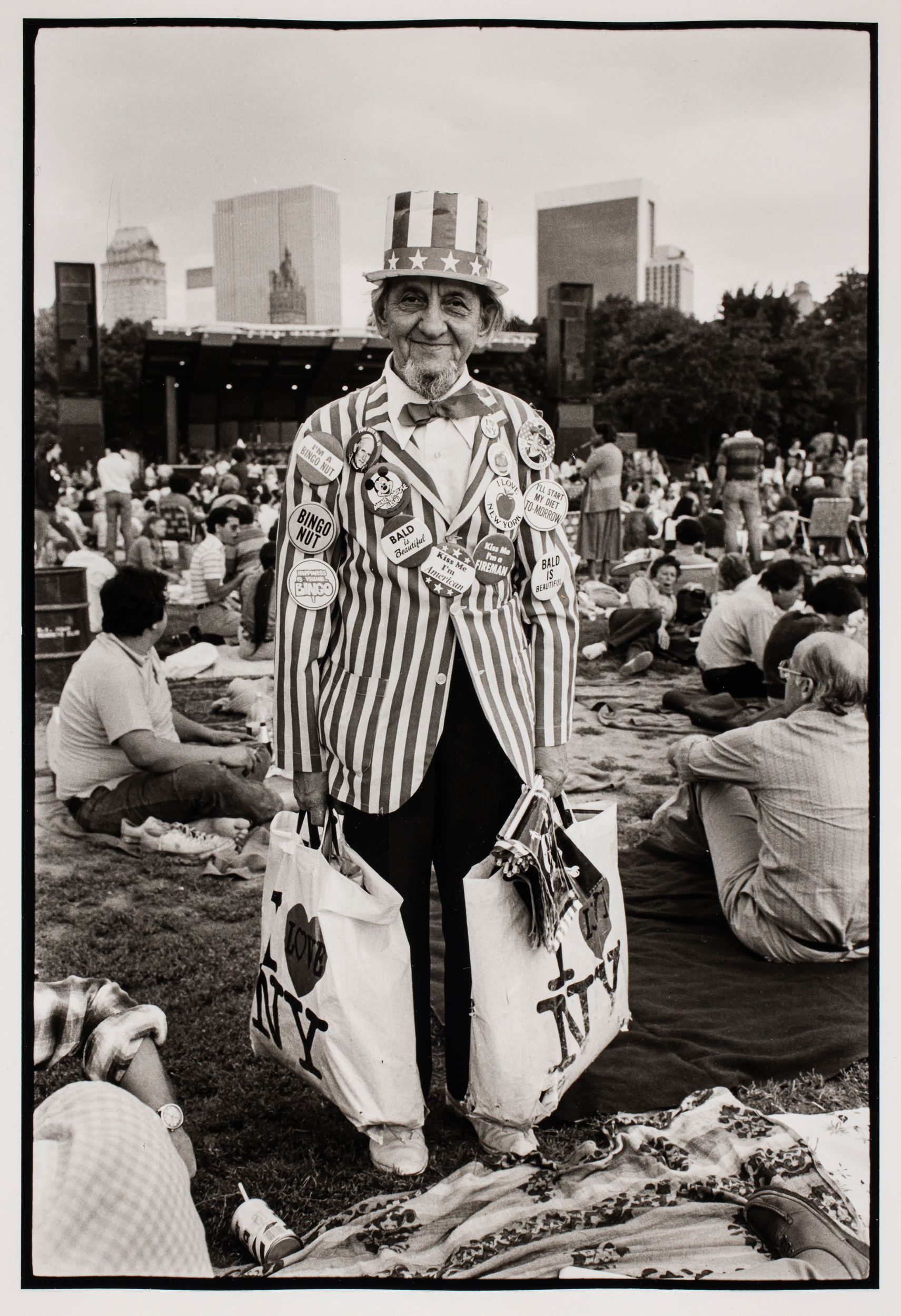 An elderly man in a patriotic outfit stands in a park holding "I Love NY" bags at an event.