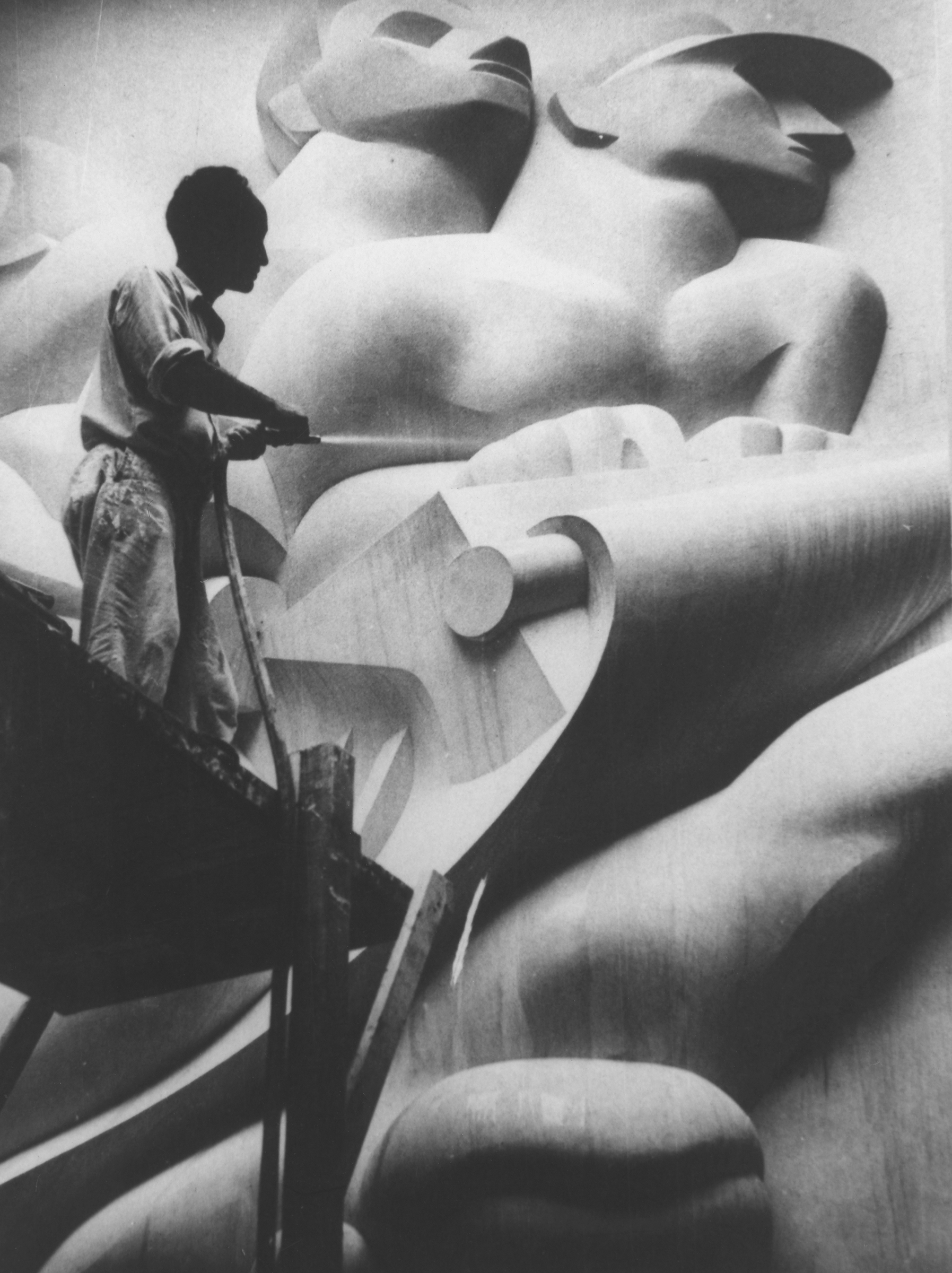 A person works on a large, abstract sculpture or mural with prominent human figures.