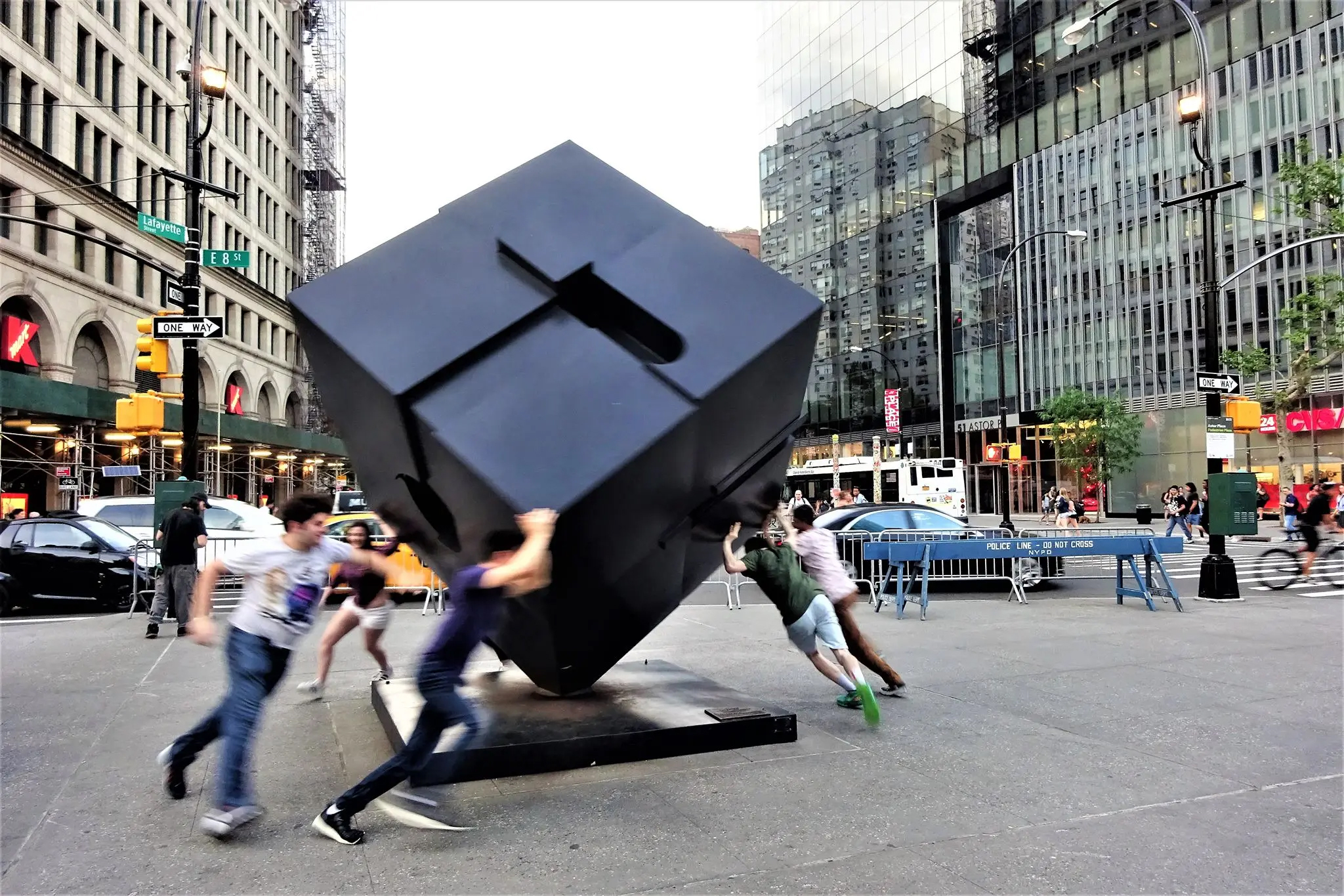 People are spinning a large black cube sculpture in a busy city intersection.