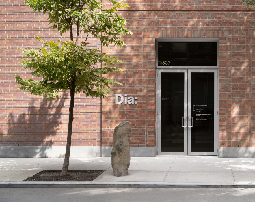 A modern brick building entrance with "Dia:" on the wall, a tree, and a standing stone outside.
