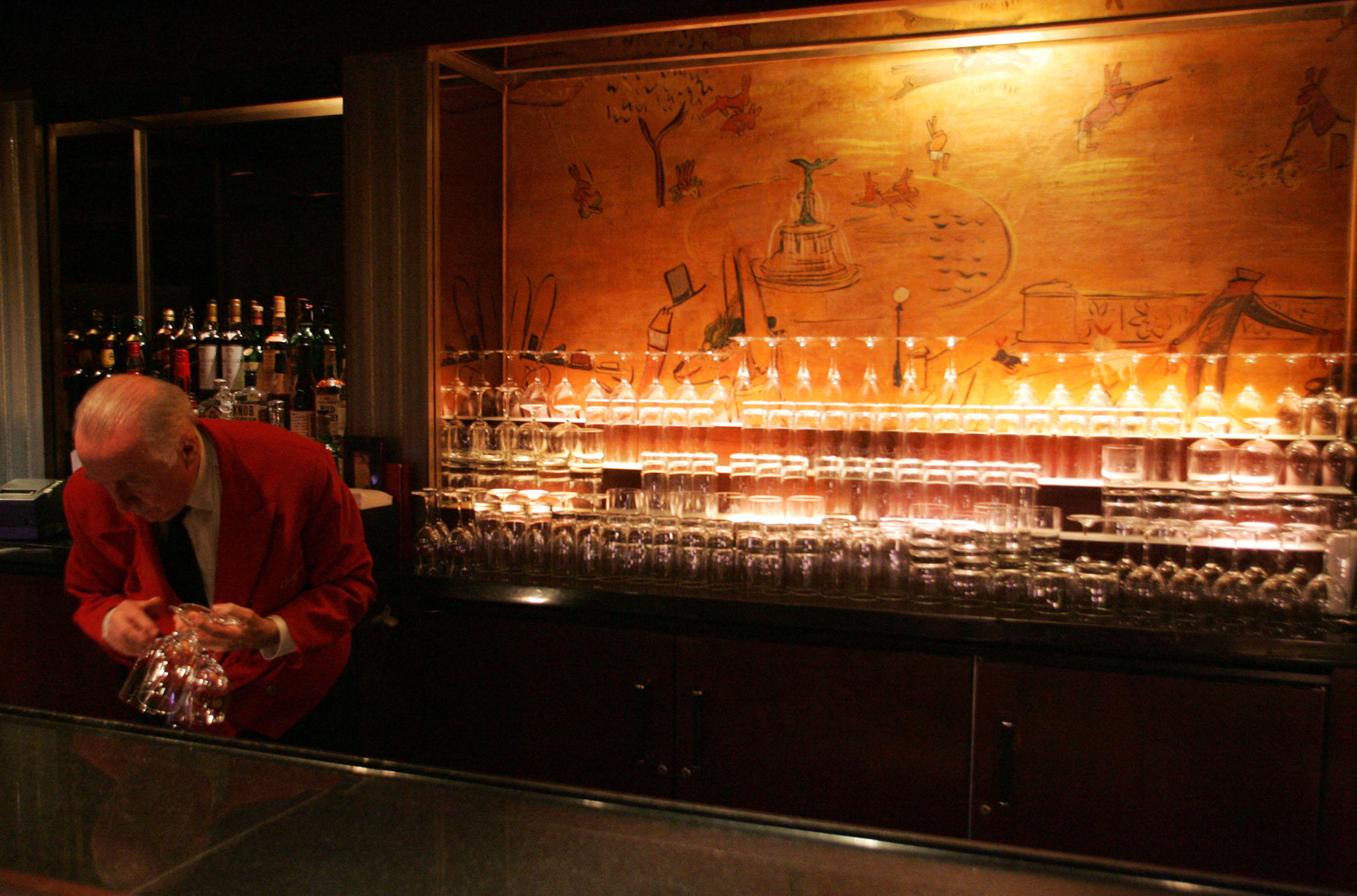 An elderly bartender in a red jacket polishes glasses behind a dimly lit bar with a mural.