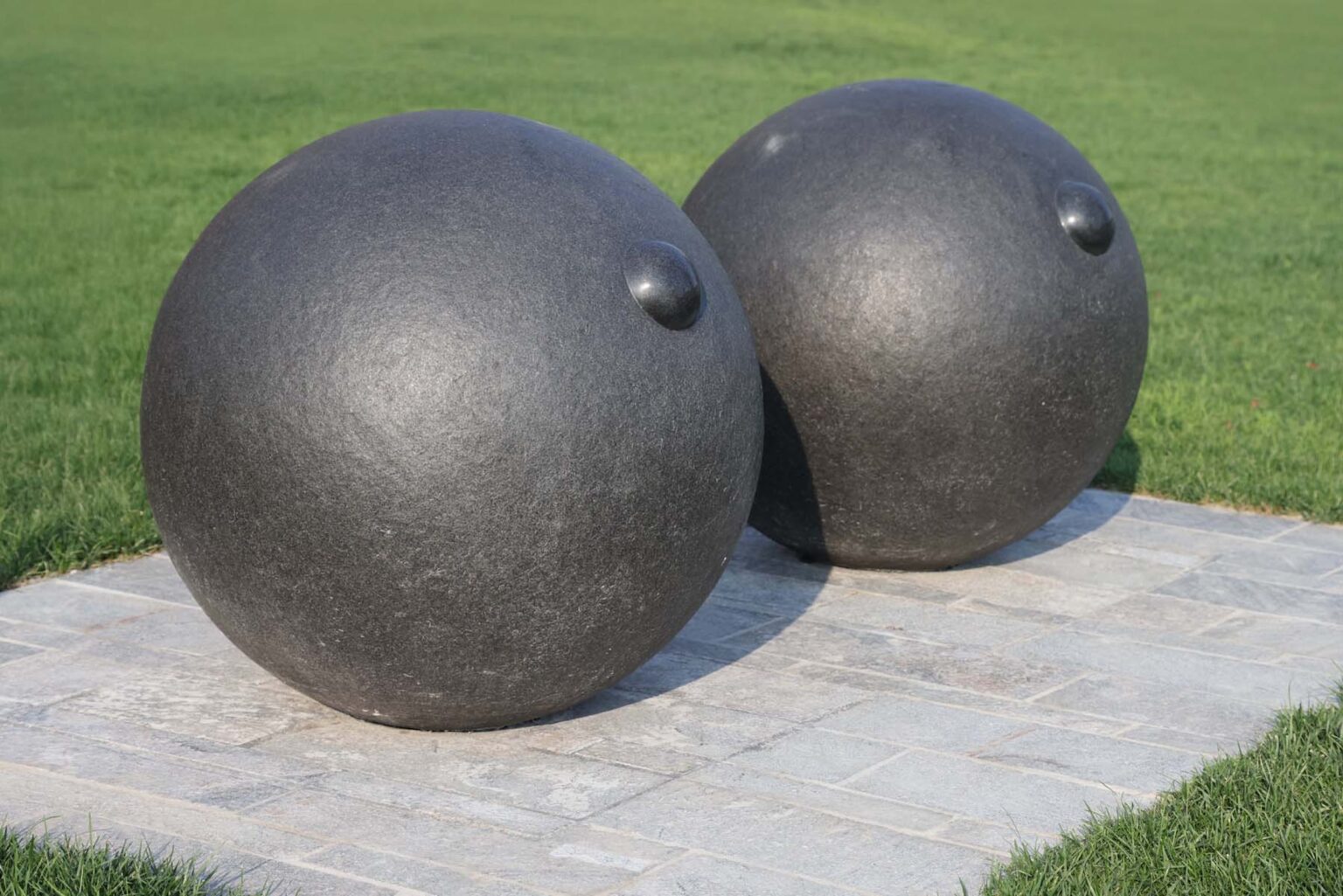 Two large, round black sculptures with small protrusions sit on a stone platform in the grass.