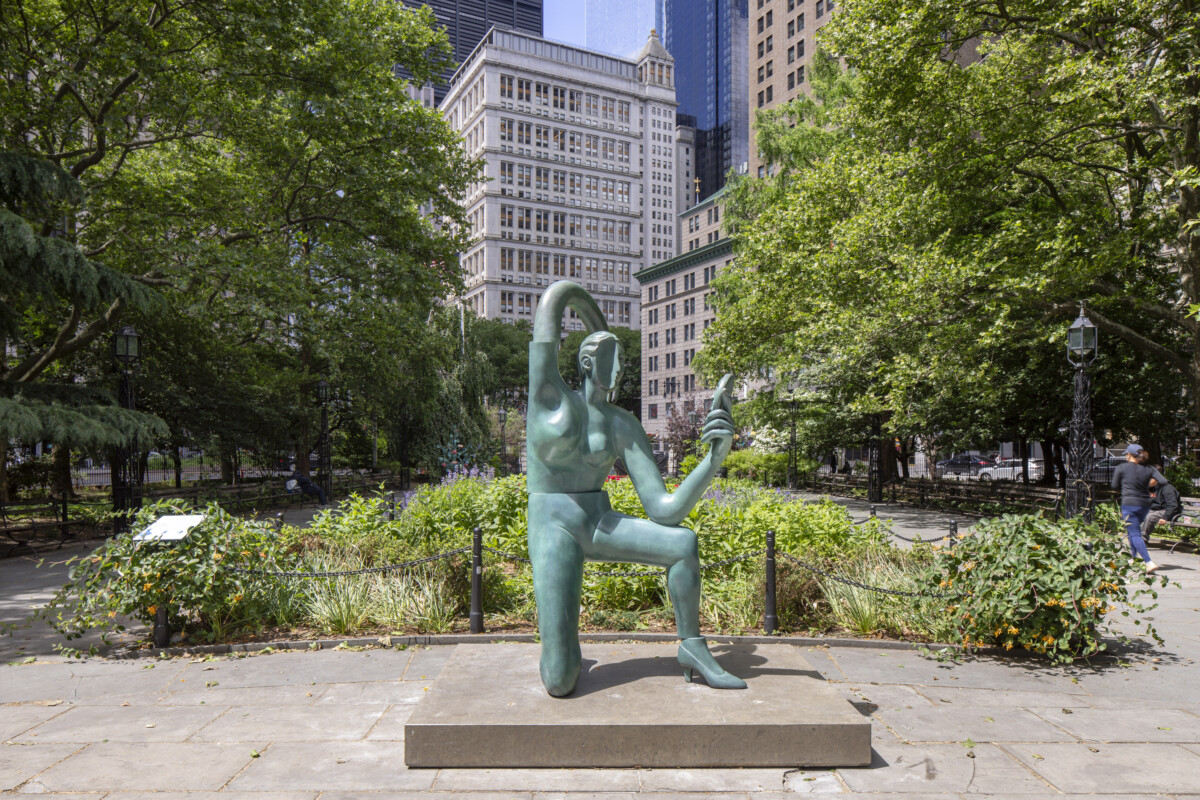 A modern abstract sculpture stands in a city park surrounded by trees and tall buildings.