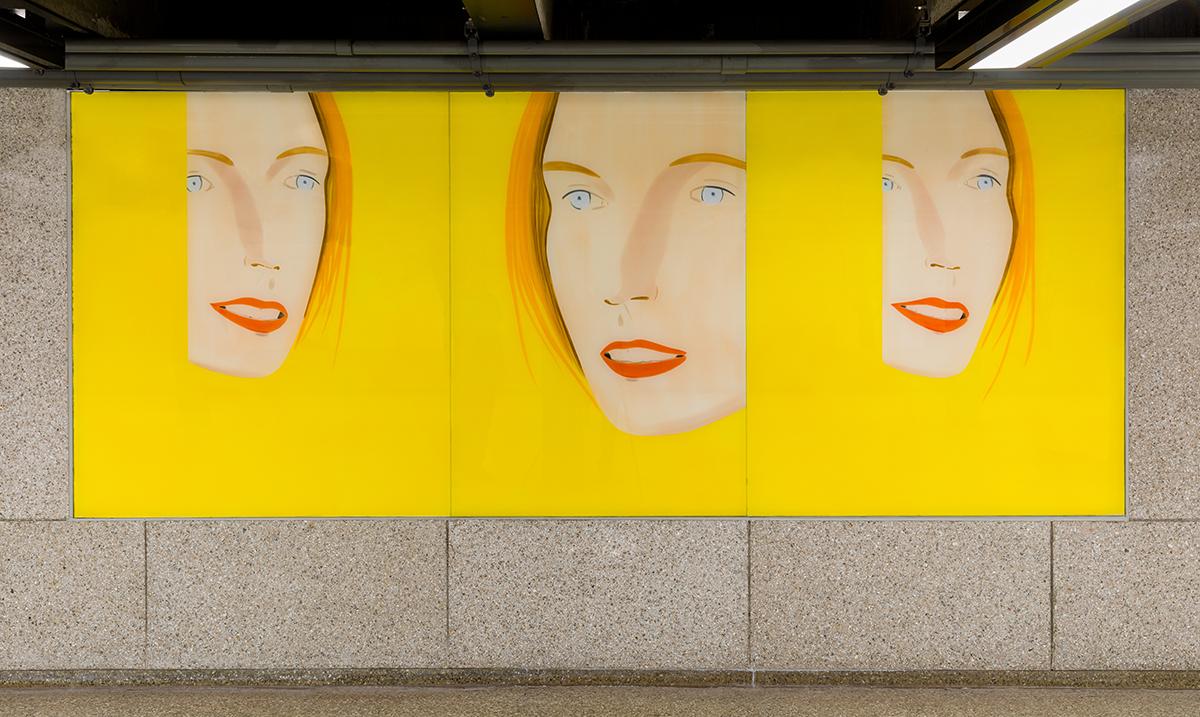 A mural with three minimalist female faces on a bright yellow background in a subway station.