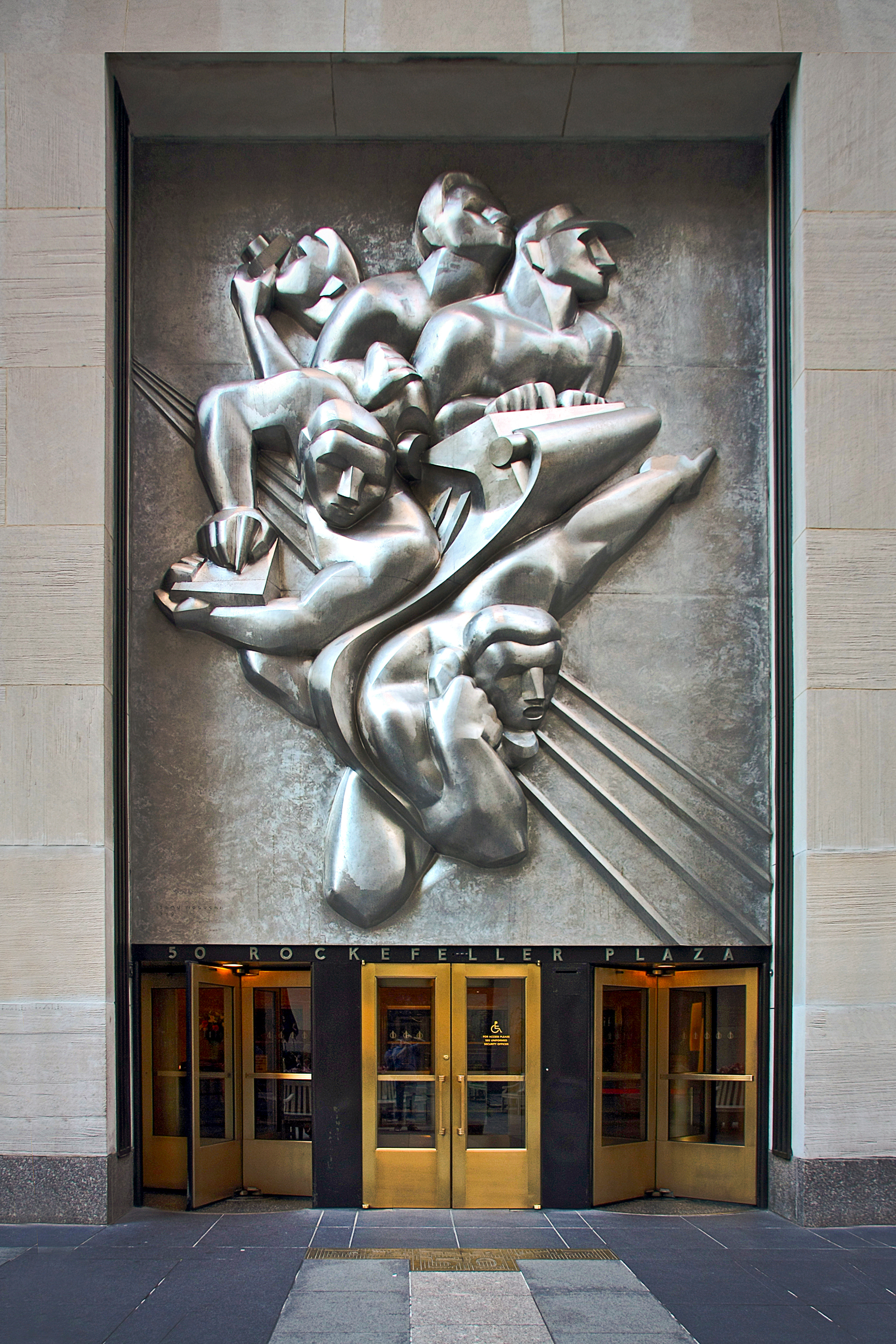 A large silver bas-relief sculpture of stylized human figures is above gold revolving doors.