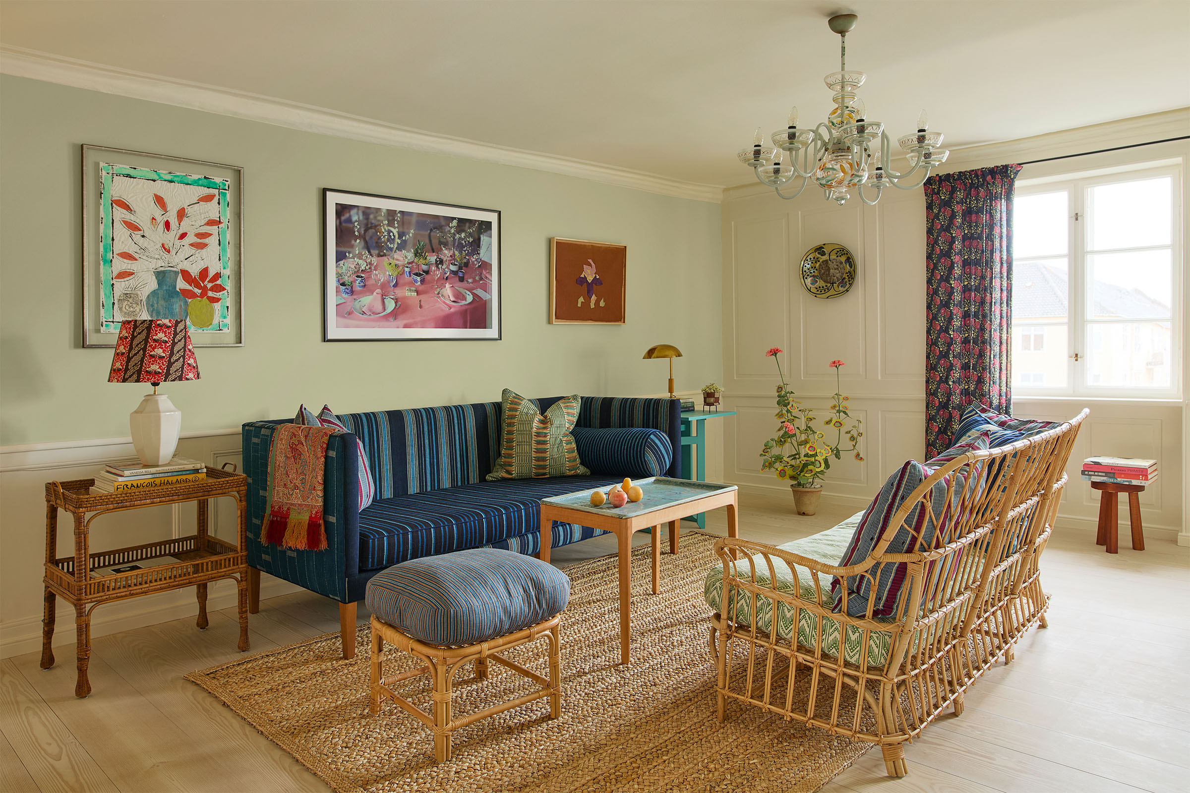 The drawing room of The Apartment. On the walls, from left, are Isabella Ducrot's Pots with Green Frame I, 2023; Lunch table, Chateau Mouton, 1977, a photograph by Peter Schlesinger; Yto Barrada’s Untitled, 2022; and a 1960s ceramic by Birger Kaipiainen. The wicker sofa and stool are by Svenskt Tenn, designed by Josef Frank. The 1950s brass table lamp and the chandelier overhead are by the Finnish designer Paavo Tynell. The fabric shade on the lamp to the left of the sofa, standing on a 1930s wicker tray table, is by Sibyl Colefax and John Fowler