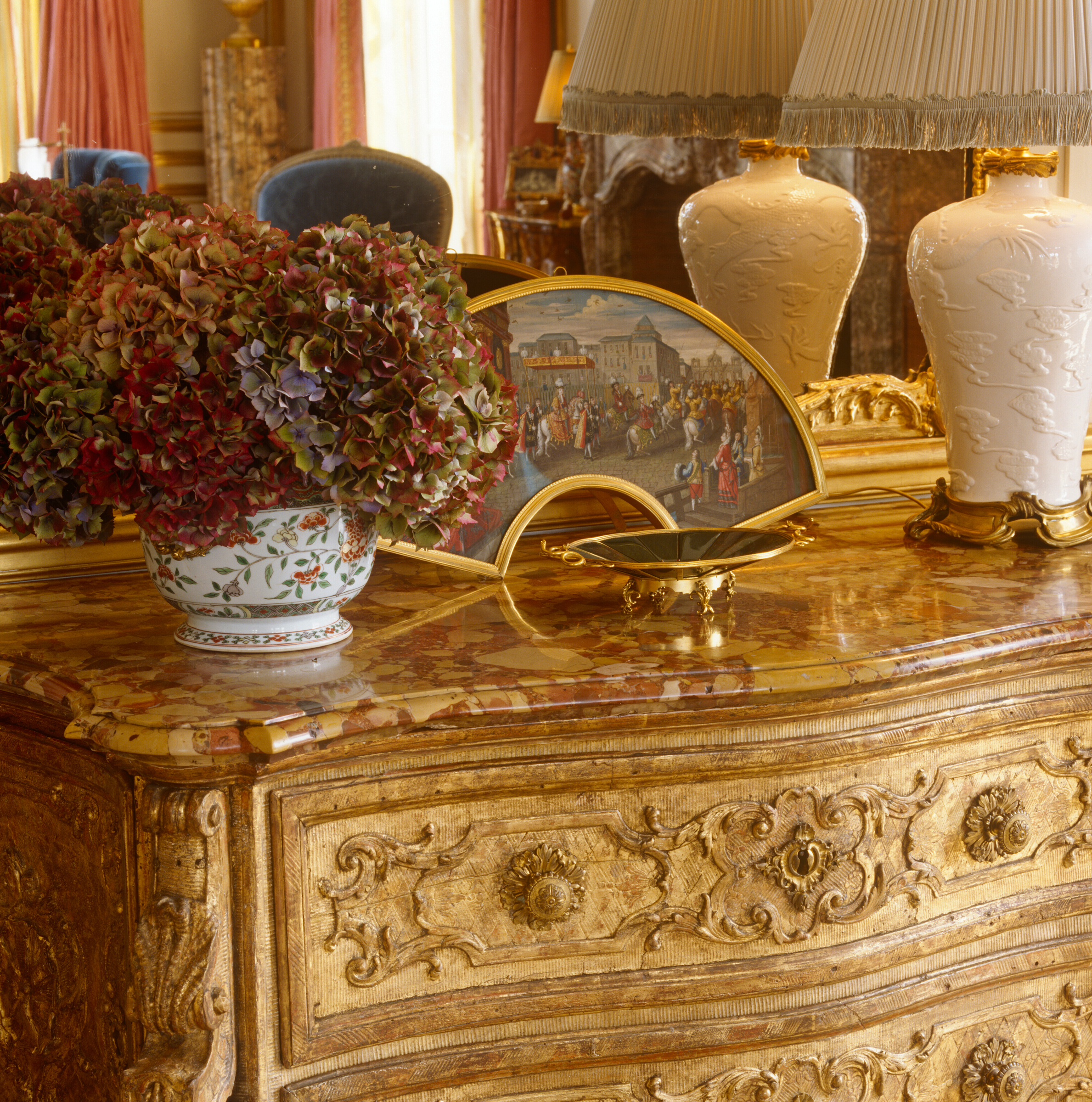 An ornate marble-topped dresser with a floral vase, lamps, and a decorative painted fan.
