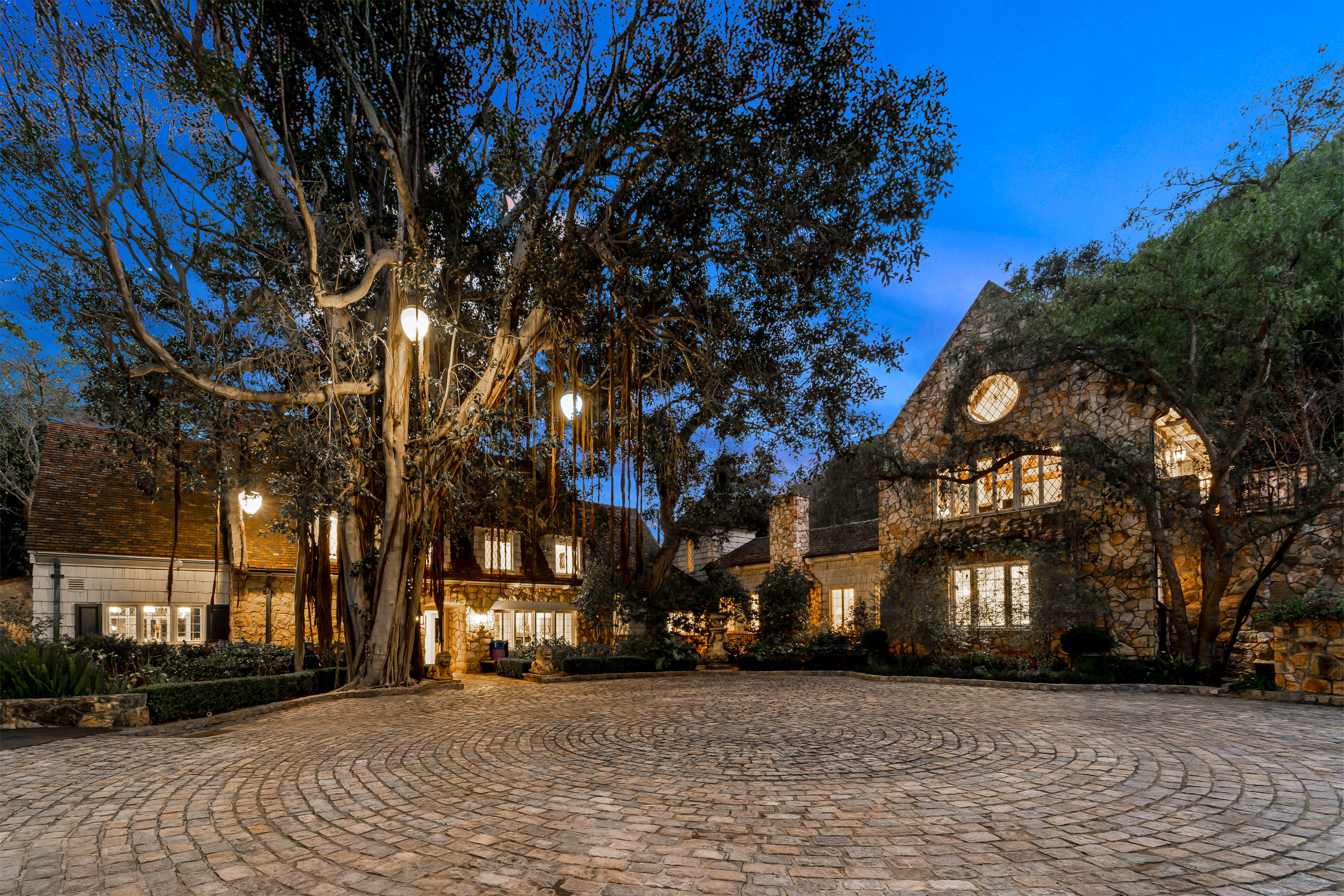 A large stone house with illuminated windows sits behind a cobblestone driveway at dusk.