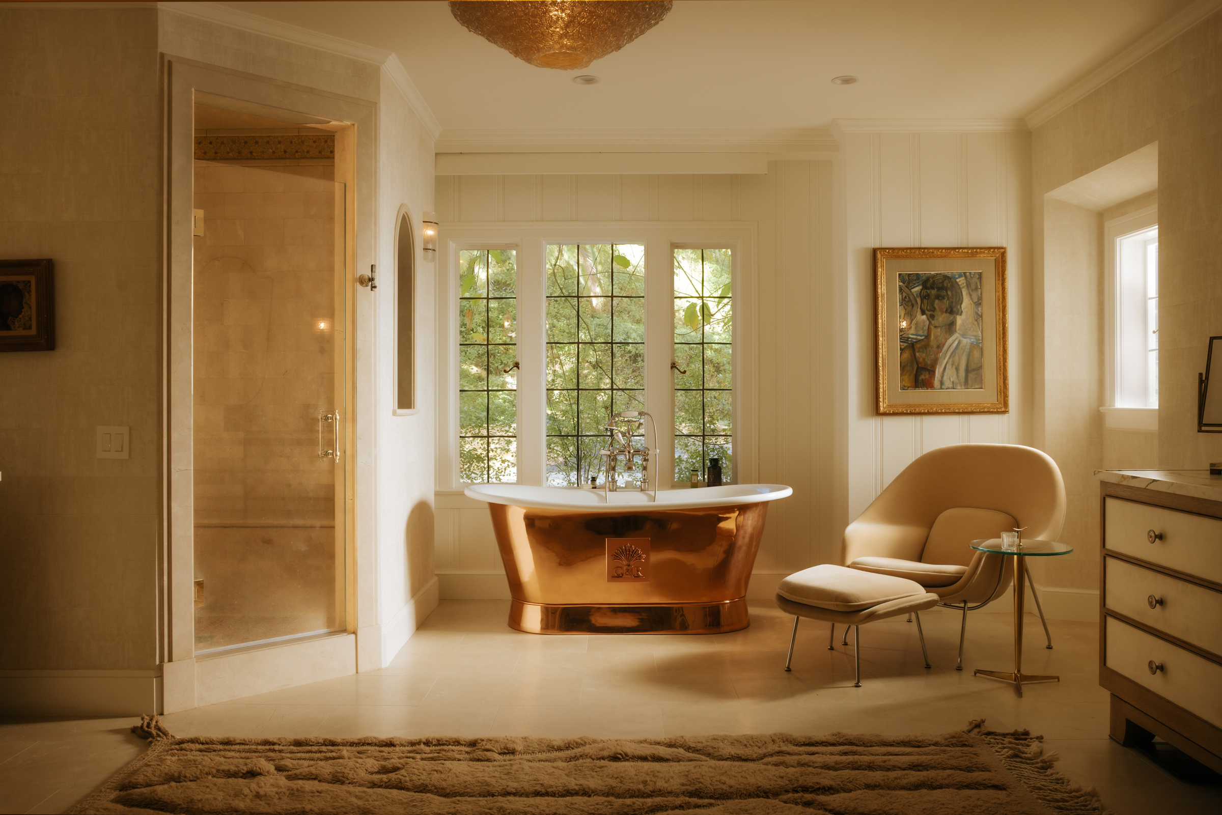 A luxurious bathroom features a copper bathtub, modern chair, glass shower, and large windows.