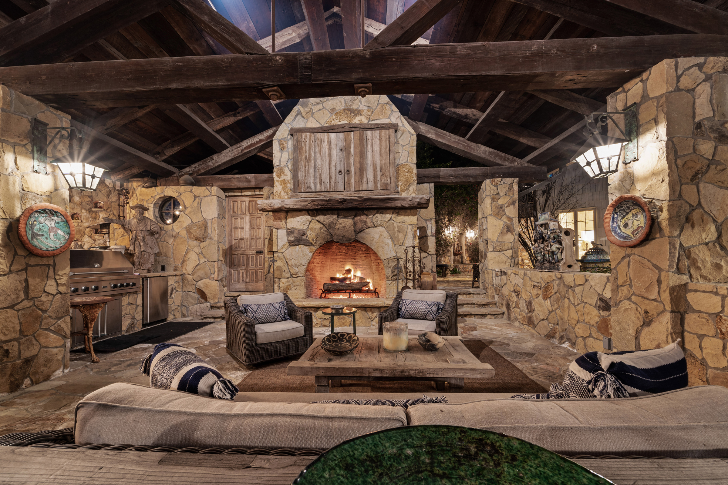 Rustic outdoor living area with stone walls, fireplace, wooden beams, and cozy seating.