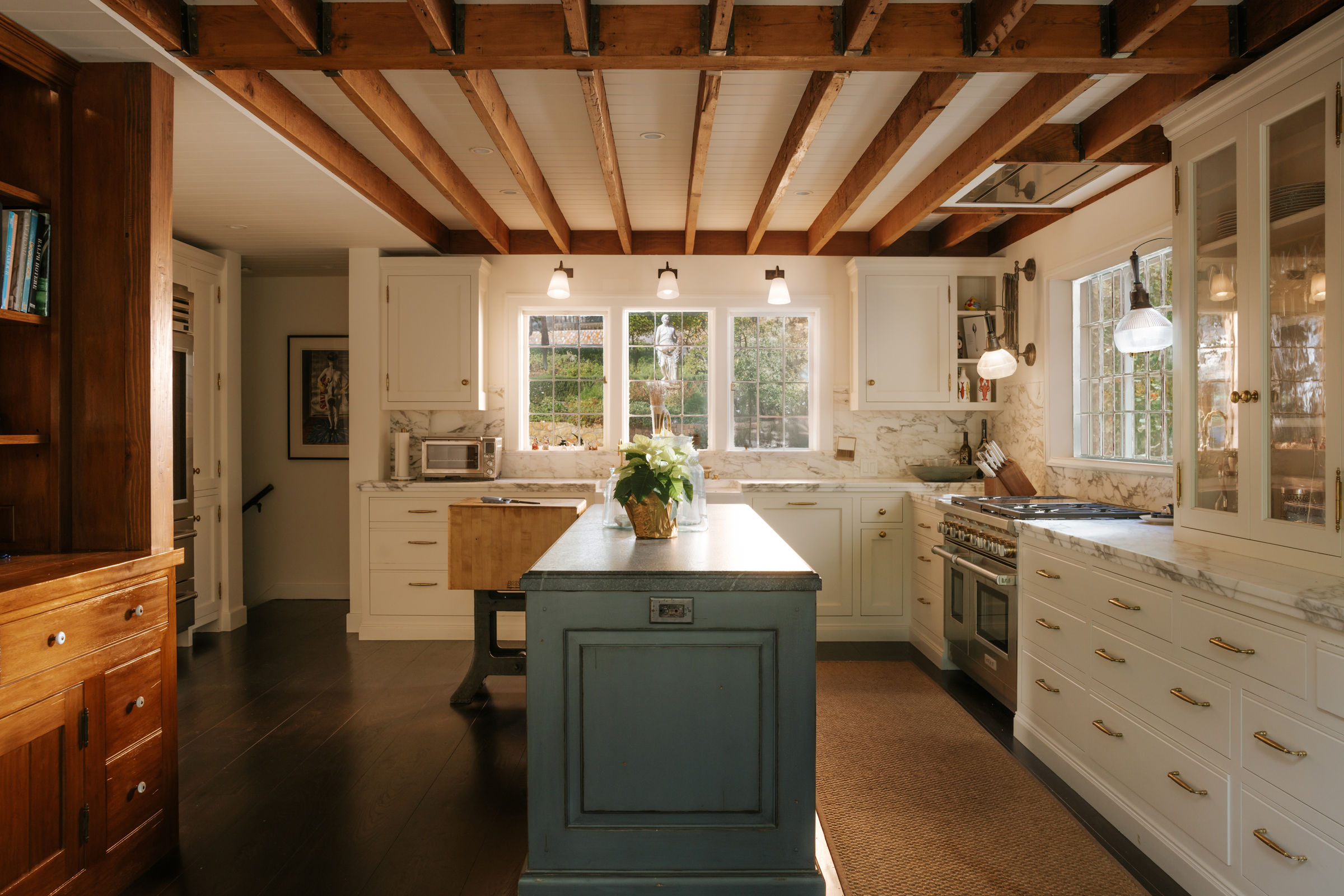 A cozy kitchen features exposed wooden beams, white cabinets, and a blue island with a plant.