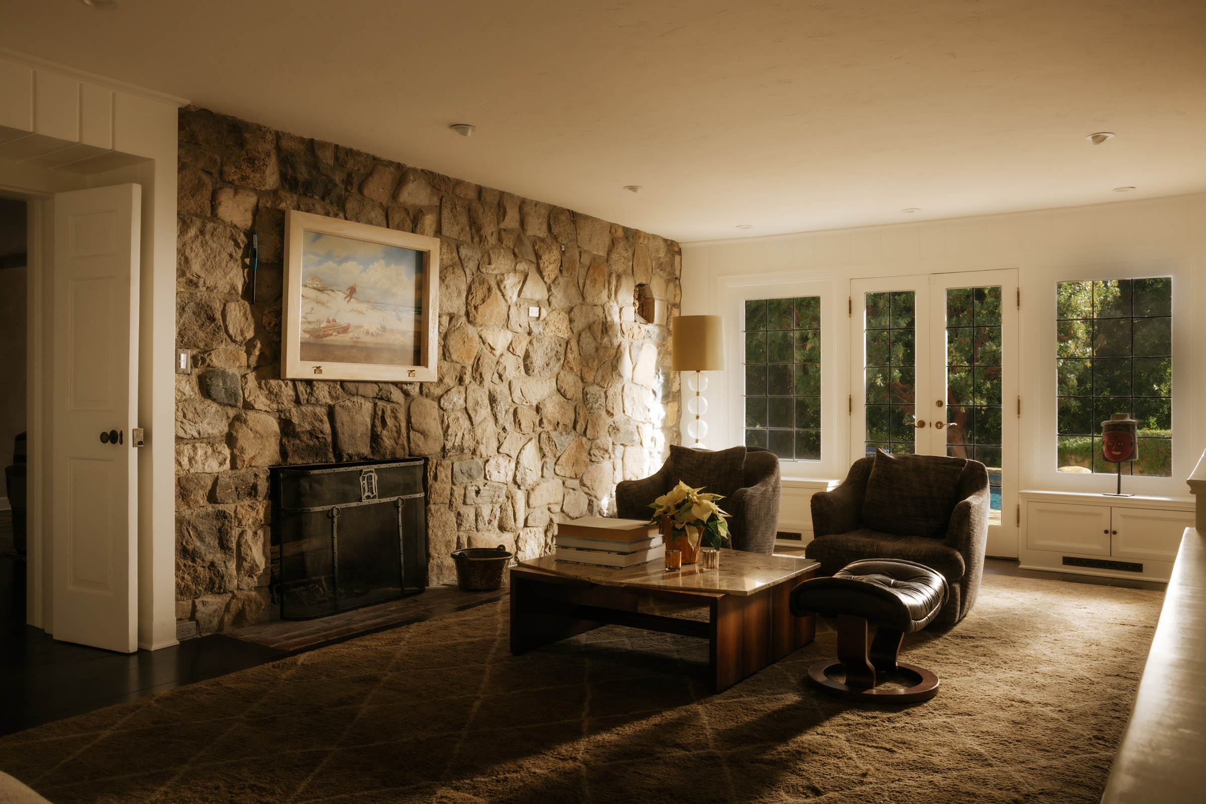 A cozy living room with stone fireplace, armchairs, coffee table, and large windows.