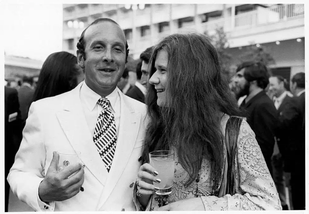 A man in a white suit and a woman with long hair smile at an outdoor social gathering.