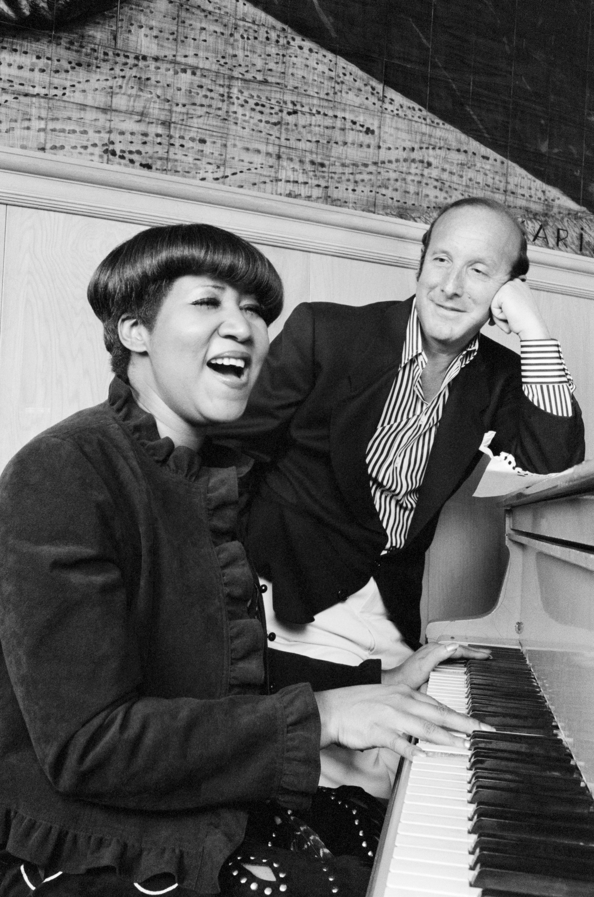 A woman is playing the piano and smiling while a man sits beside her, looking at her happily.