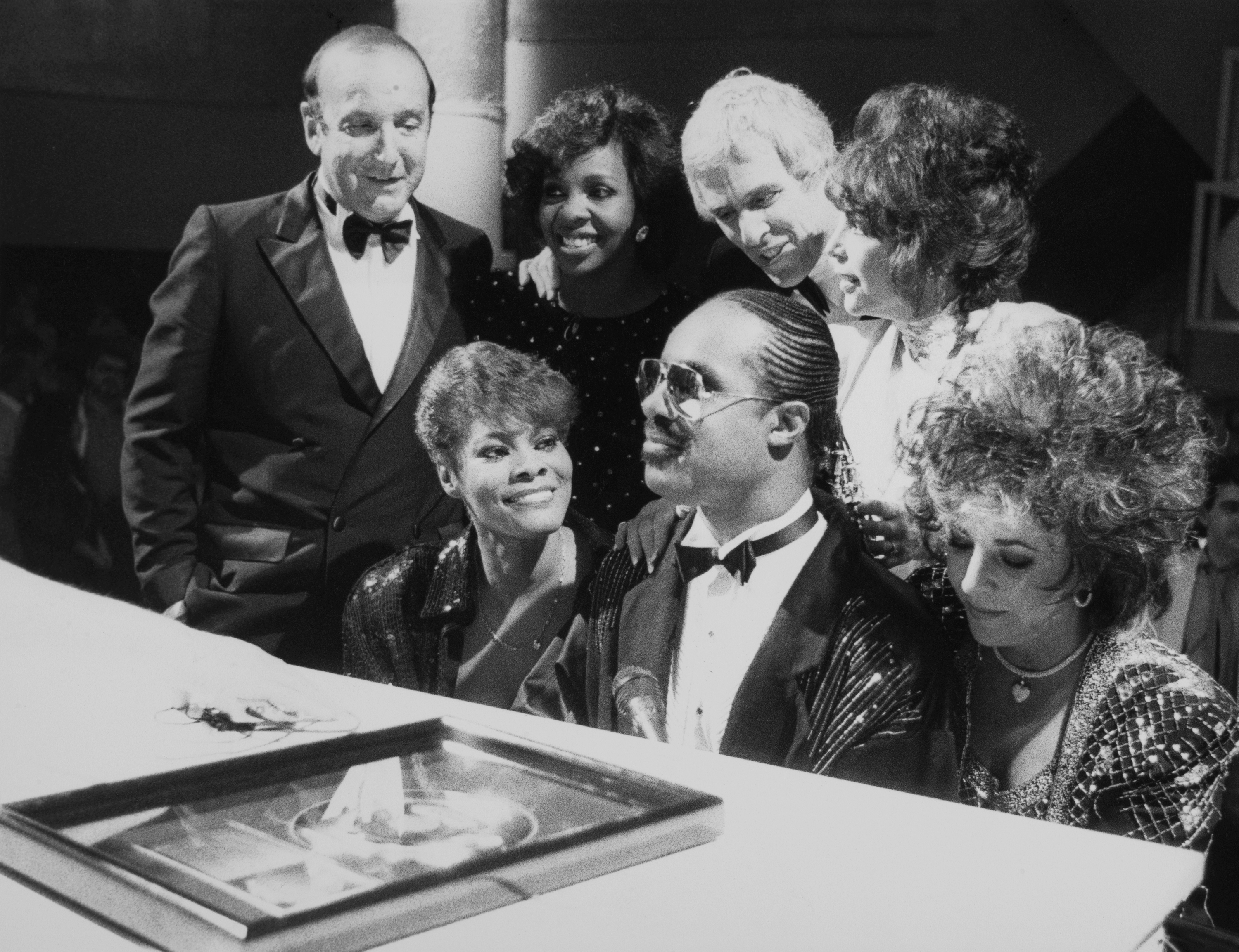 A group of elegantly dressed people are gathered around a piano, smiling and posing together.