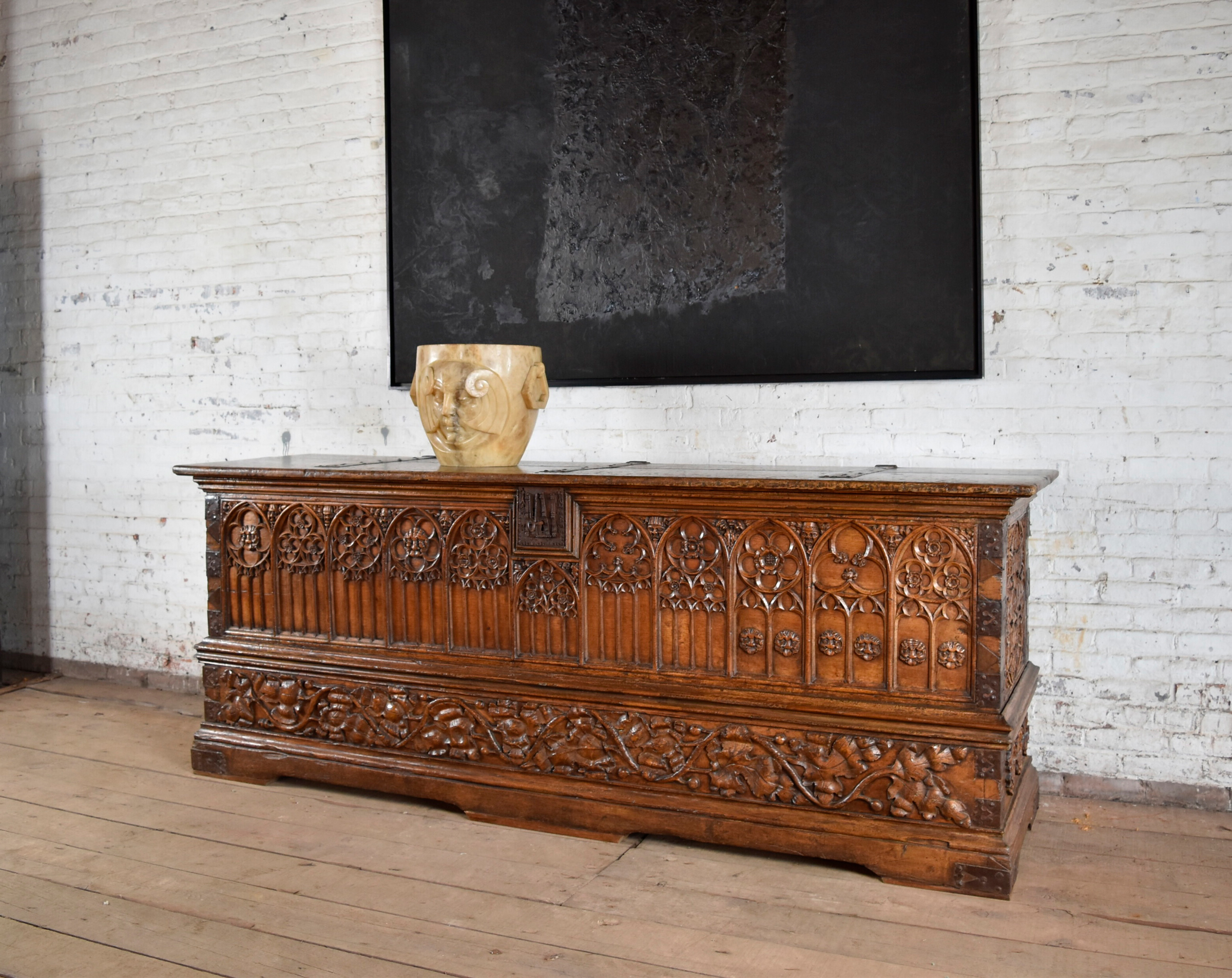 An ornate wooden chest sits against a white brick wall, with a sculpture and dark artwork above.